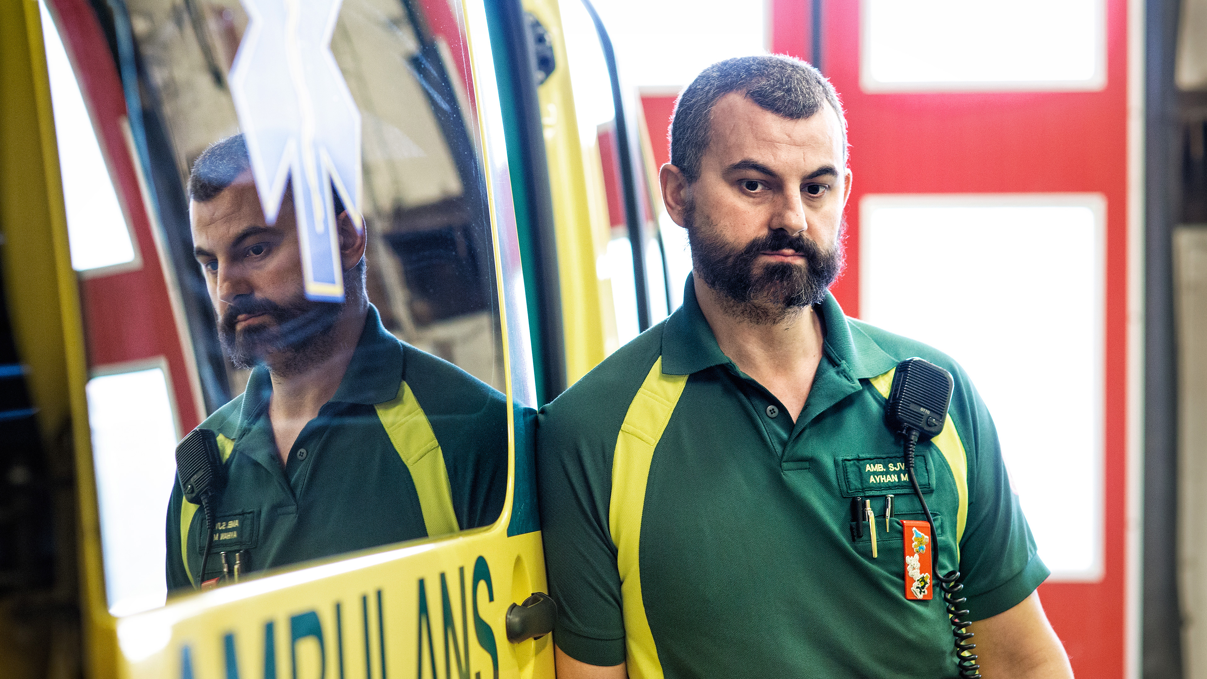 En ambulanssjukvårdare i grön och gul uniform står vid en ambulans med en radio på skjortan, reflekterad i fönstret.