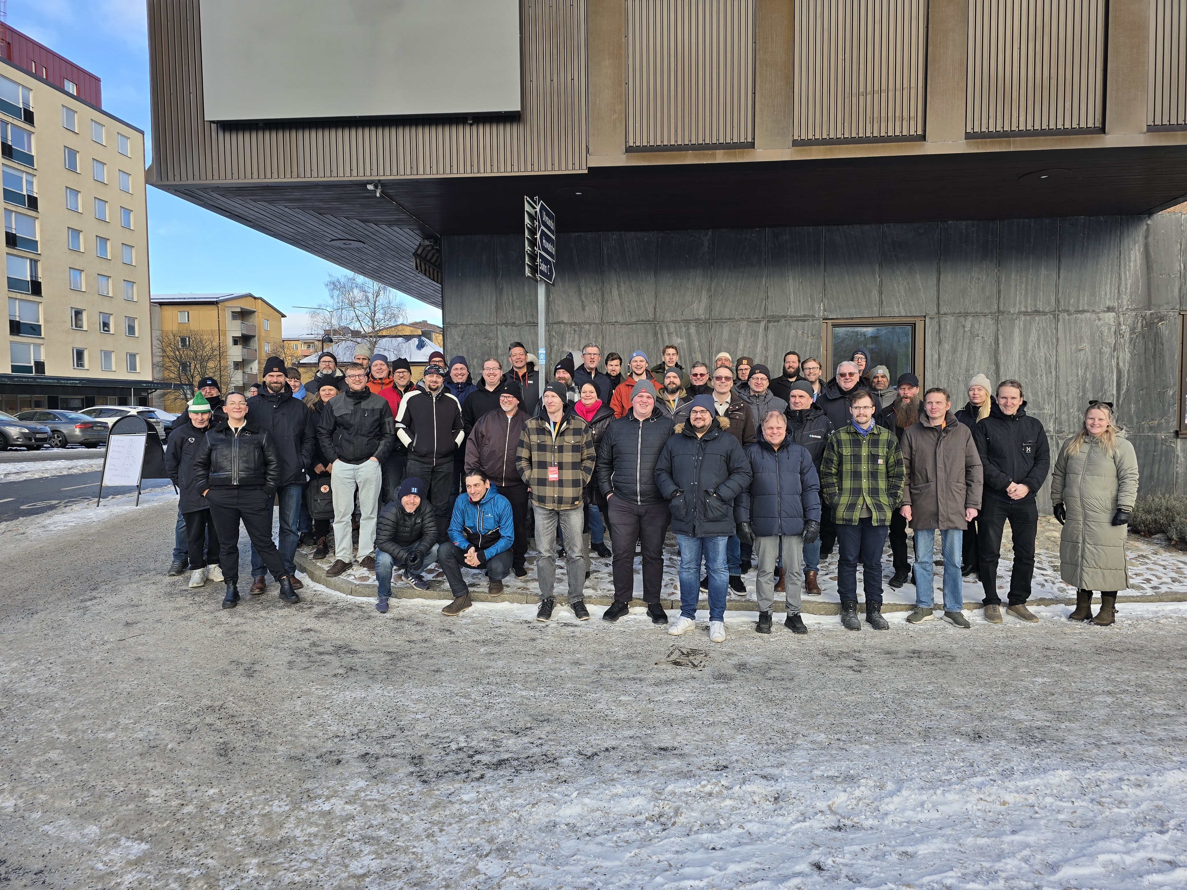 En grupp på cirka 50 personer står utanför en modern byggnad på en snötäckt dag; de flesta har vinterkläder på sig.