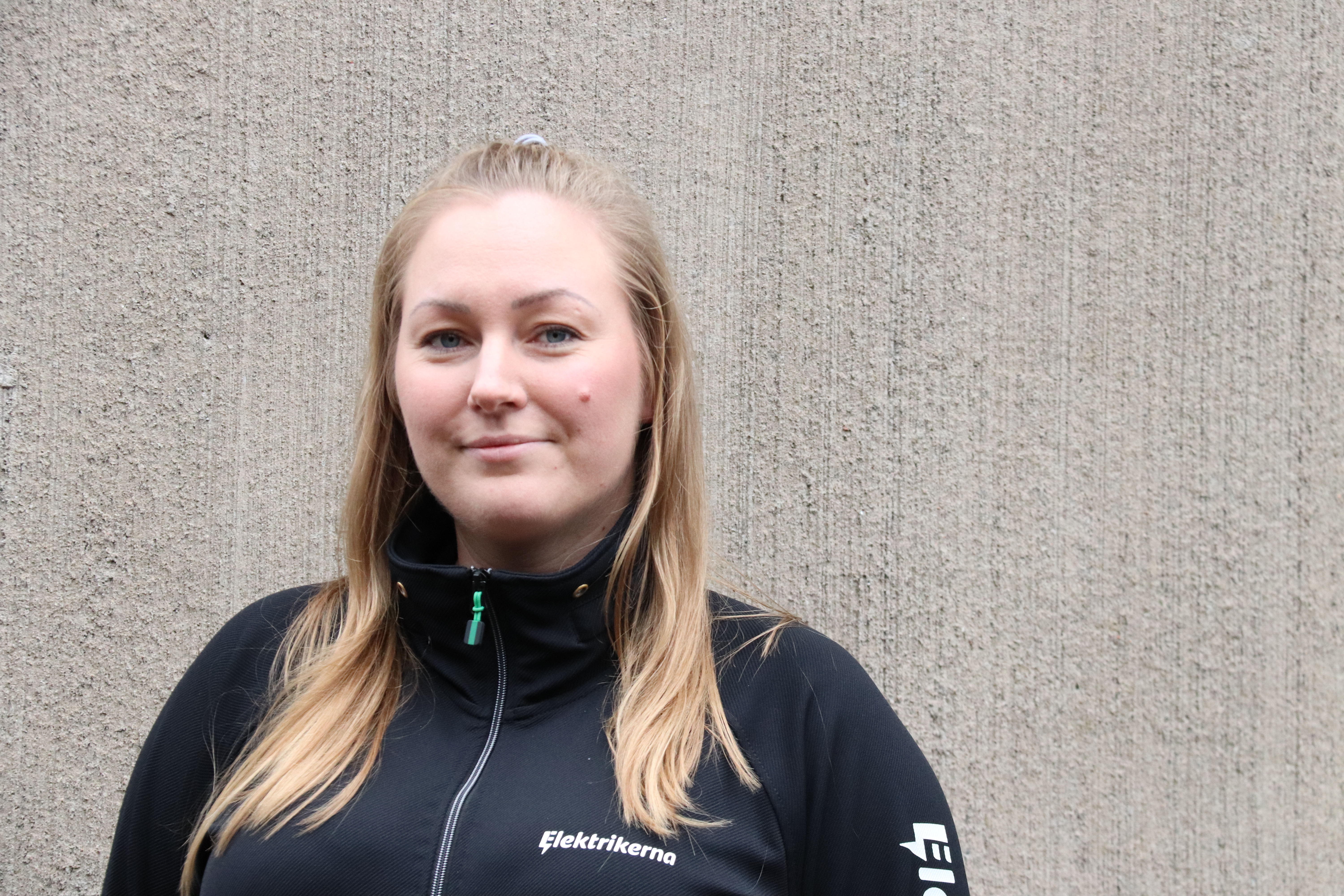 Ung kvinna med långt blont hår och svart jacka står framför en betongvägg.