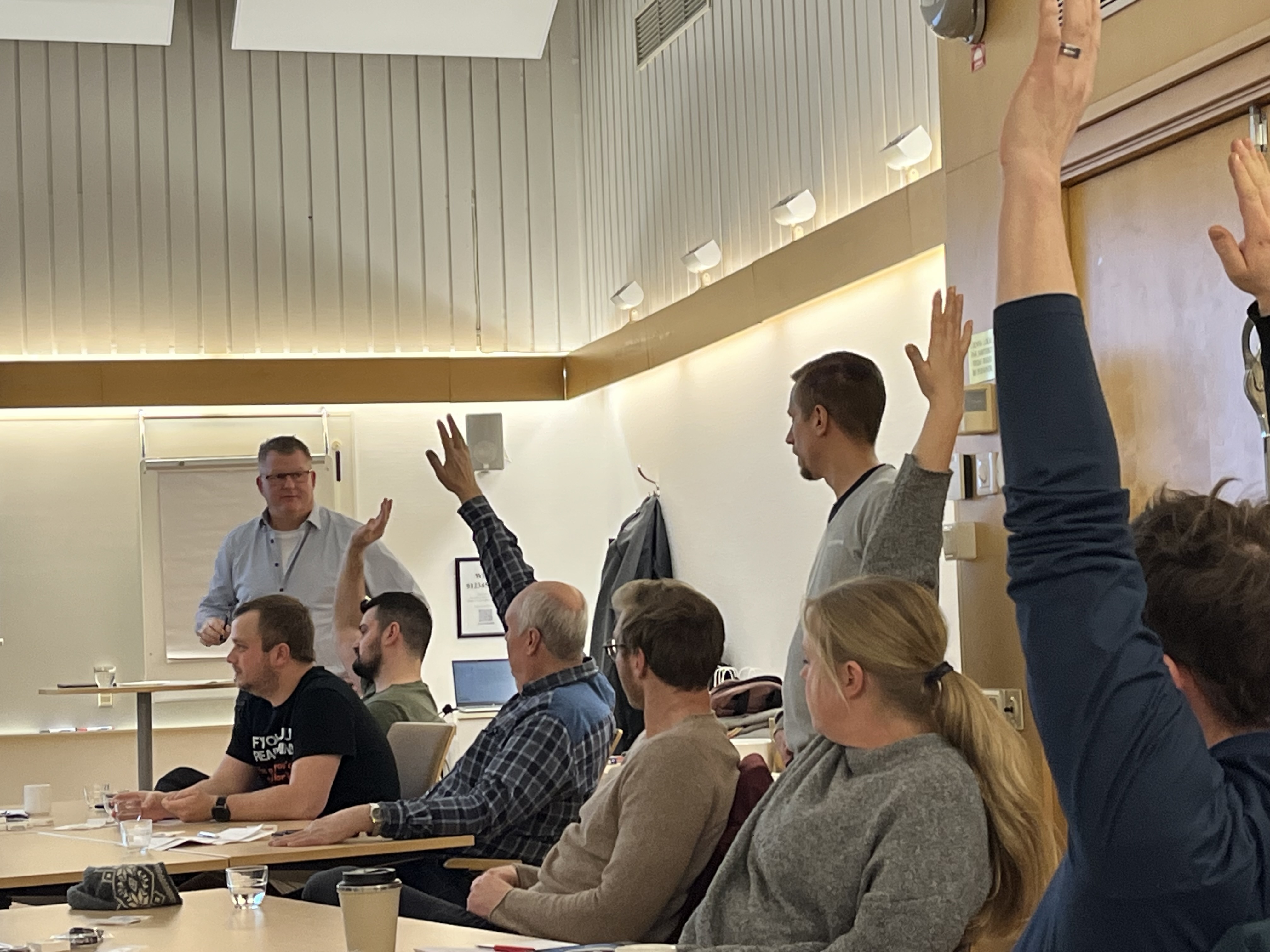 En grupp personer sitter vid bord i ett mötesrum, några räcker upp händerna medan en man står längst fram och talar.