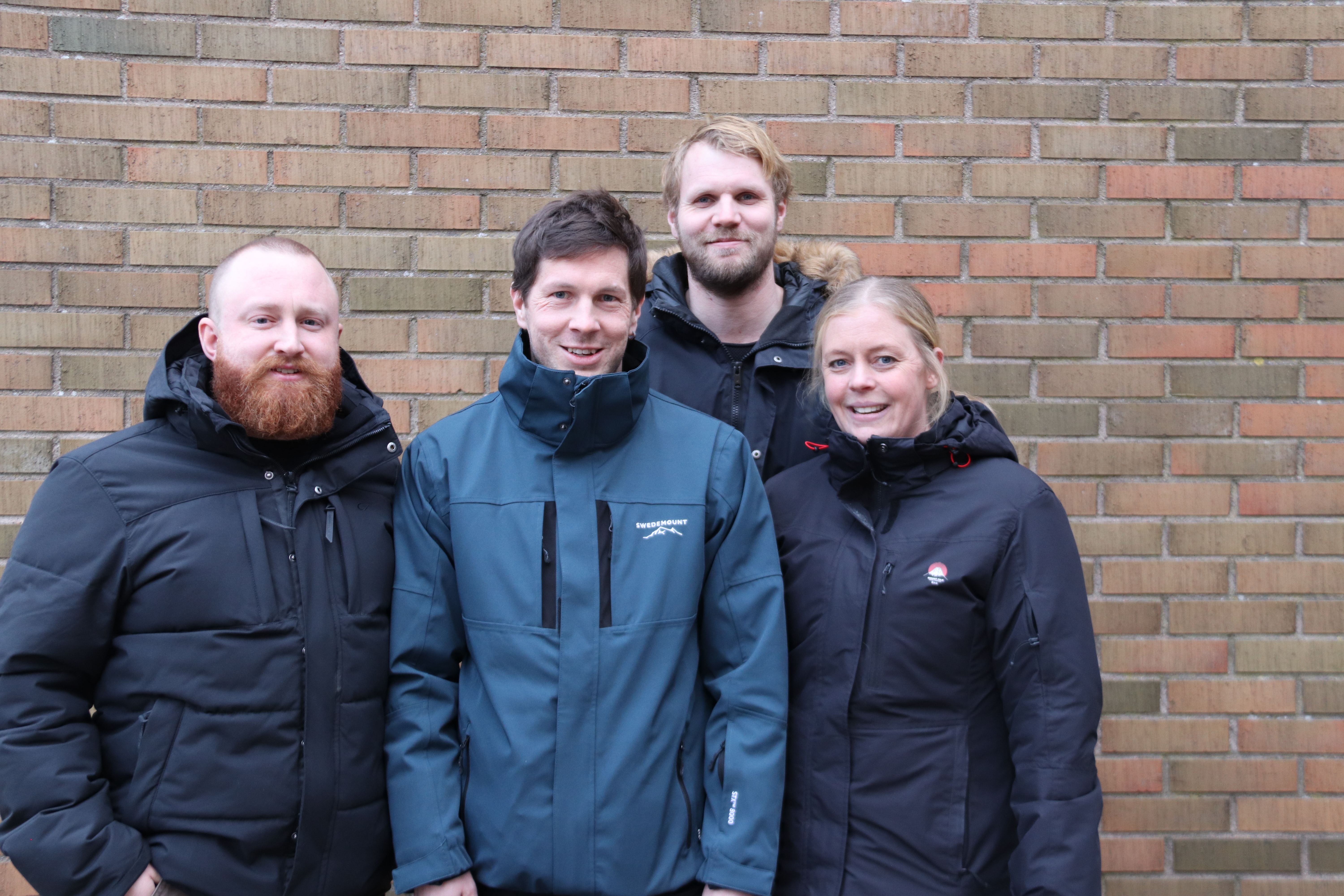 Fyra vuxna med vinterjackor står framför en tegelvägg och poserar för ett gruppfoto.