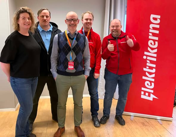 Fem personer står inomhus bredvid en röd banderoll med texten "Elektrikerna"; två har röda jackor, en gör tummen upp.