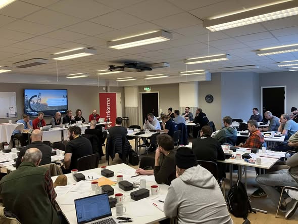 Personer sitter vid bord i ett konferensrum och deltar i möte eller workshop. En panel talar framme vid en projektorduk och banner. Papper och bärbara datorer syns på borden.