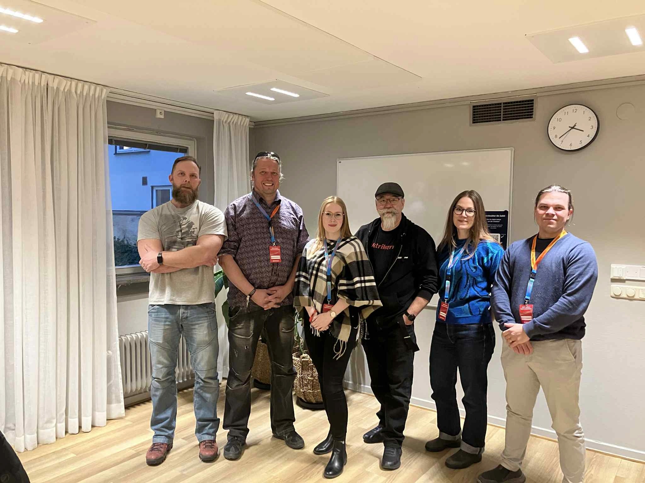 Sex personer står i ett mötesrum och poserar för en gruppbild. I bakgrunden syns en whiteboard, en klocka och ett fönster.