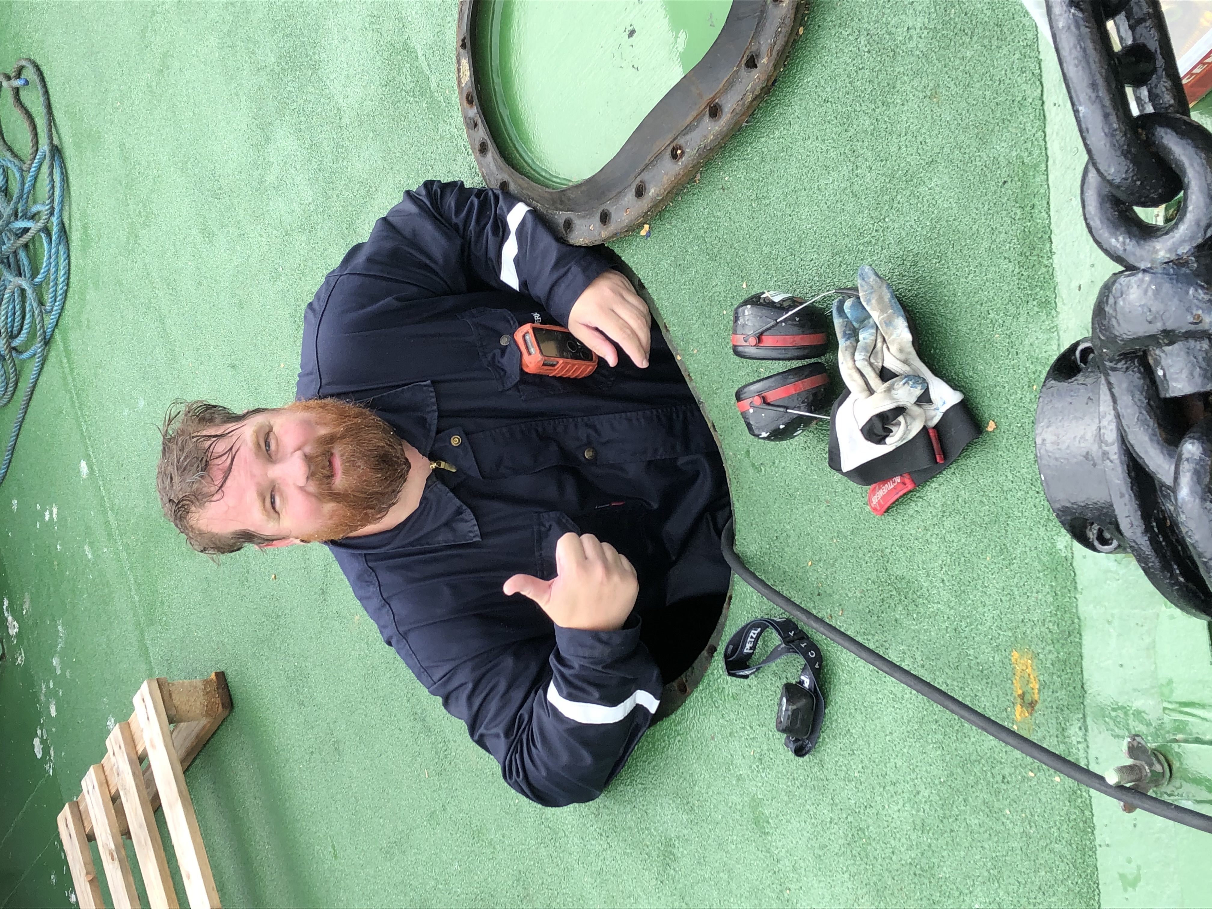 En skäggig man i arbetskläder ger en tumme upp och kommer upp ur en lucka på ett grönt däck, med skyddsutrustning och handskar nära.