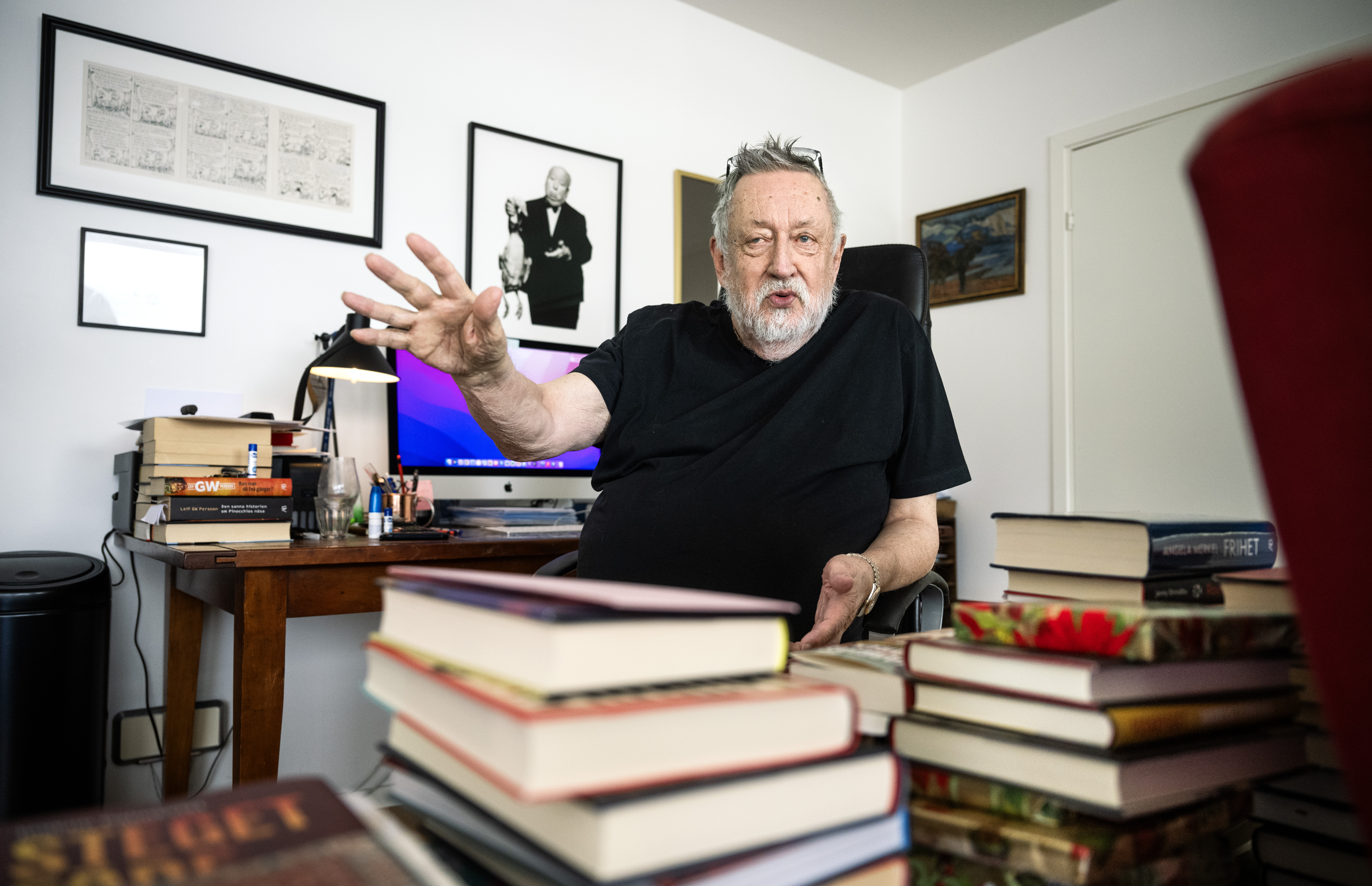 En äldre man med skägg sitter i ett hemmakontor och gör en gest med ena handen. Det finns bokstaplar framför honom, datorutrustning och inramade foton bakom honom.