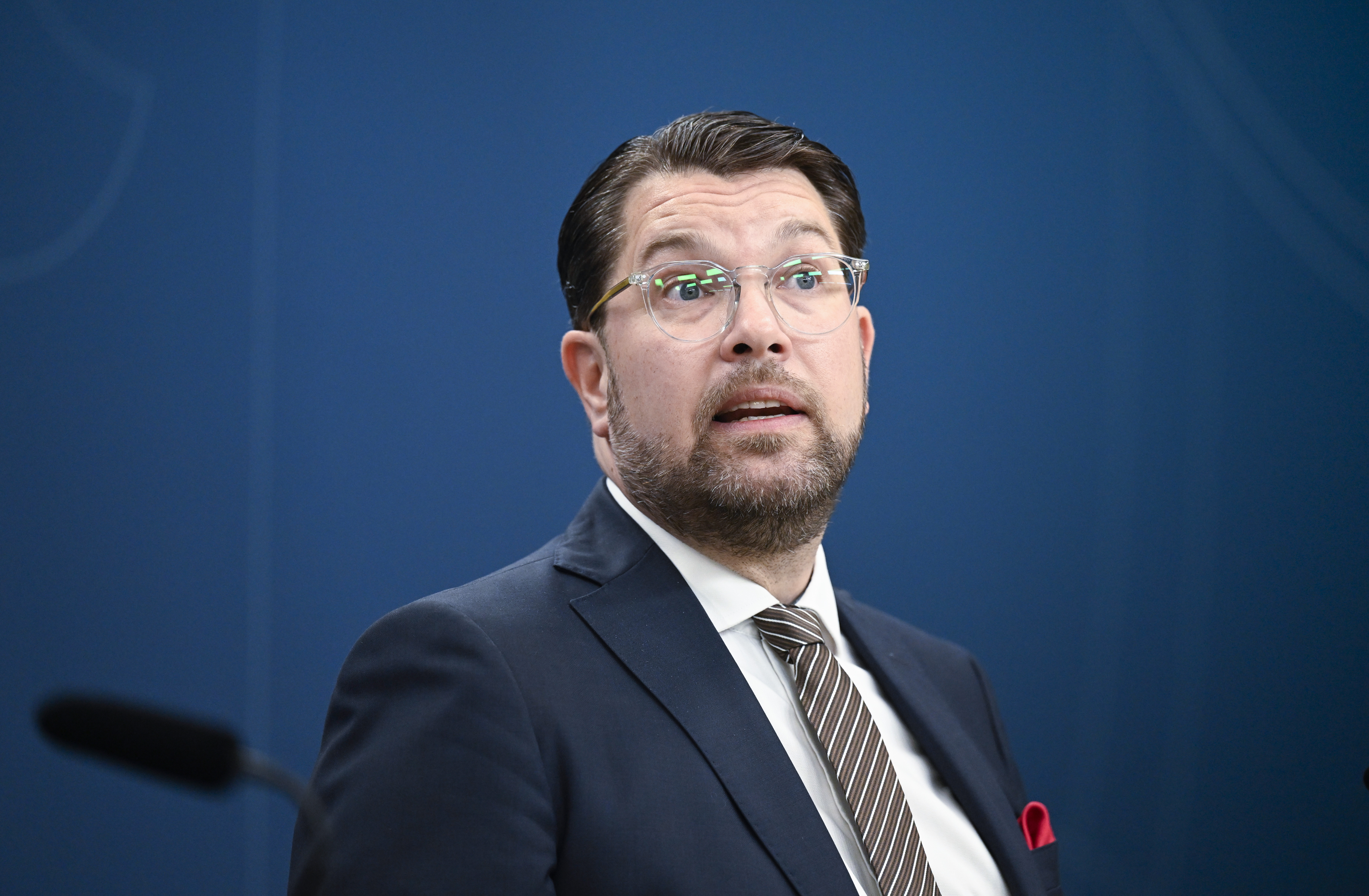 En man i kostym och slips talar vid en talarstol mot en blå bakgrund.