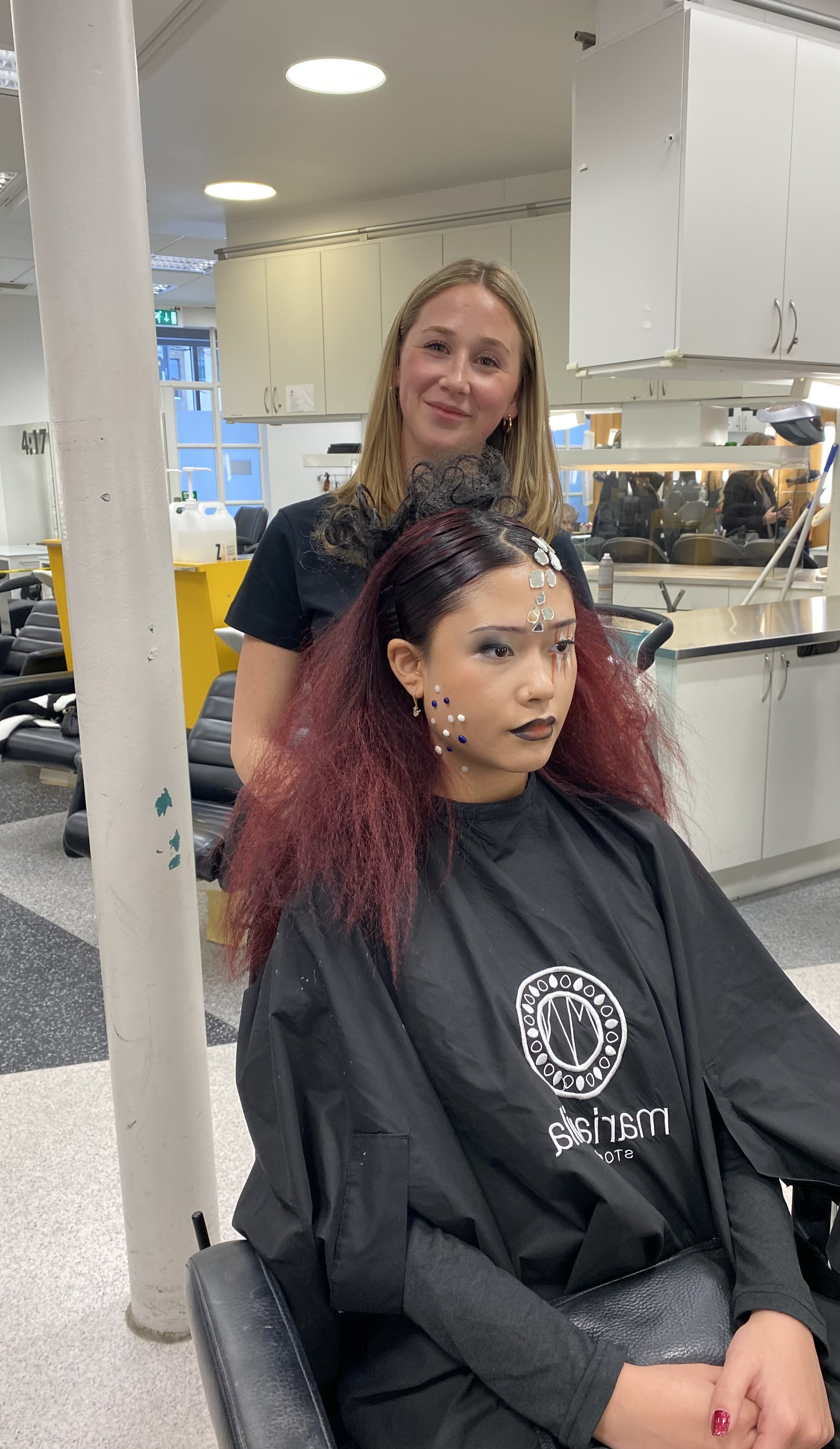 En frisör står bakom en sittande modell med mörk sminkning, mörkrött hår och dekorativa stenar i ansiktet i en salong.