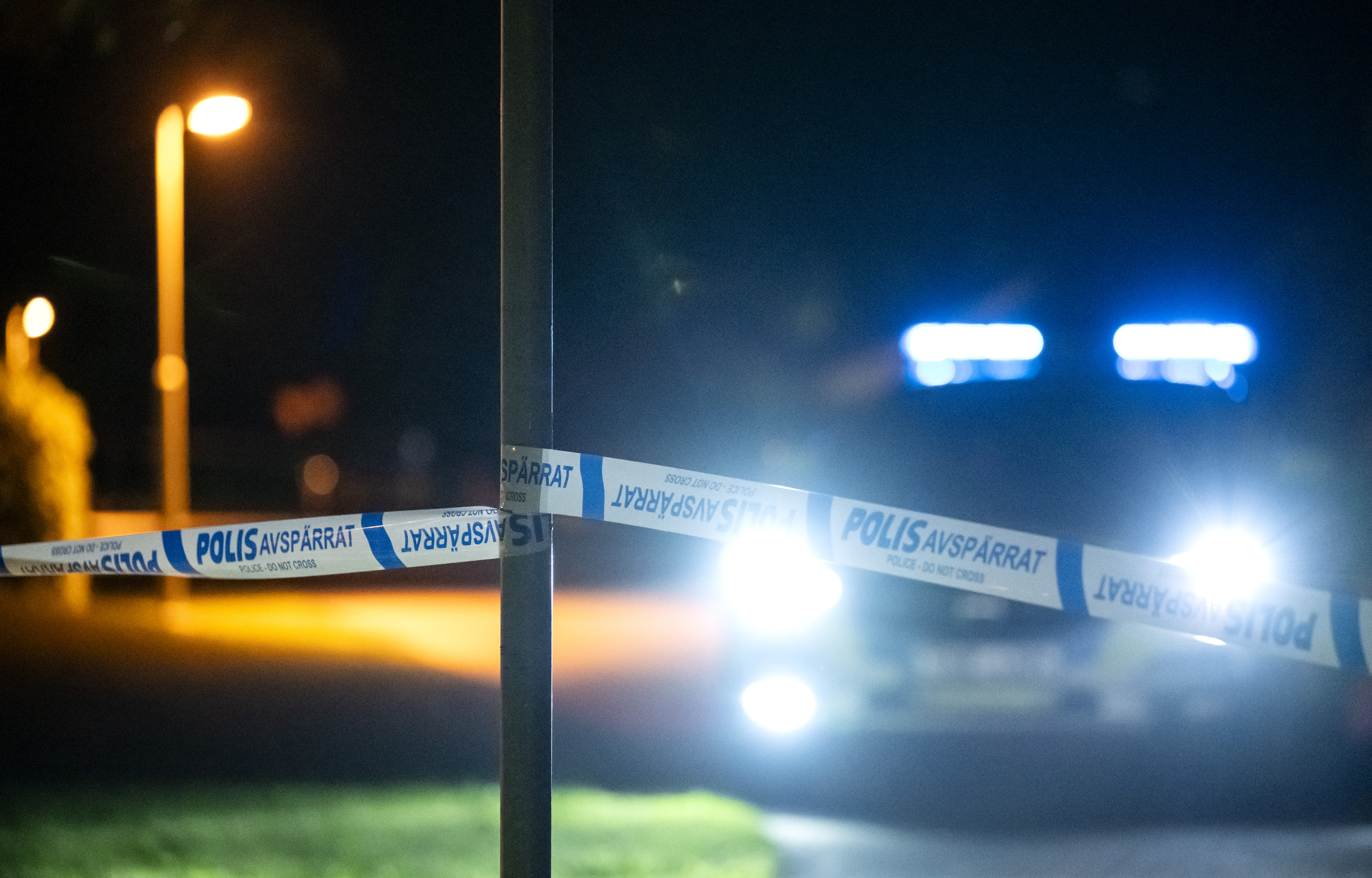 Polistejp med texten "Polis Avspärrat" spärrar av ett område på natten, en polisbil syns med tända strålkastare och blåljus.