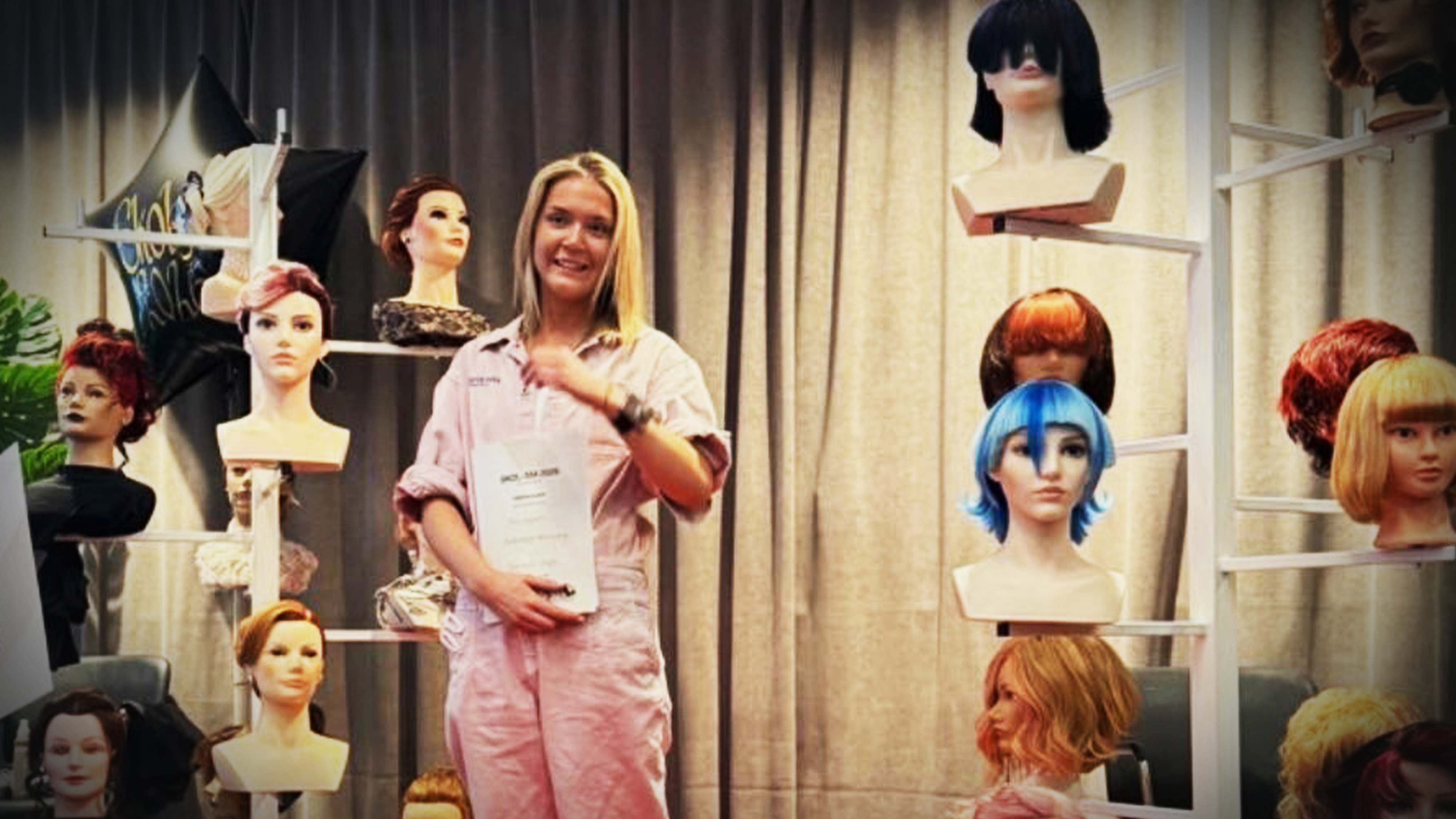 En kvinna i rosa overall står bland provhuvuden med peruker i olika stilar och färger och håller i ett häfte.