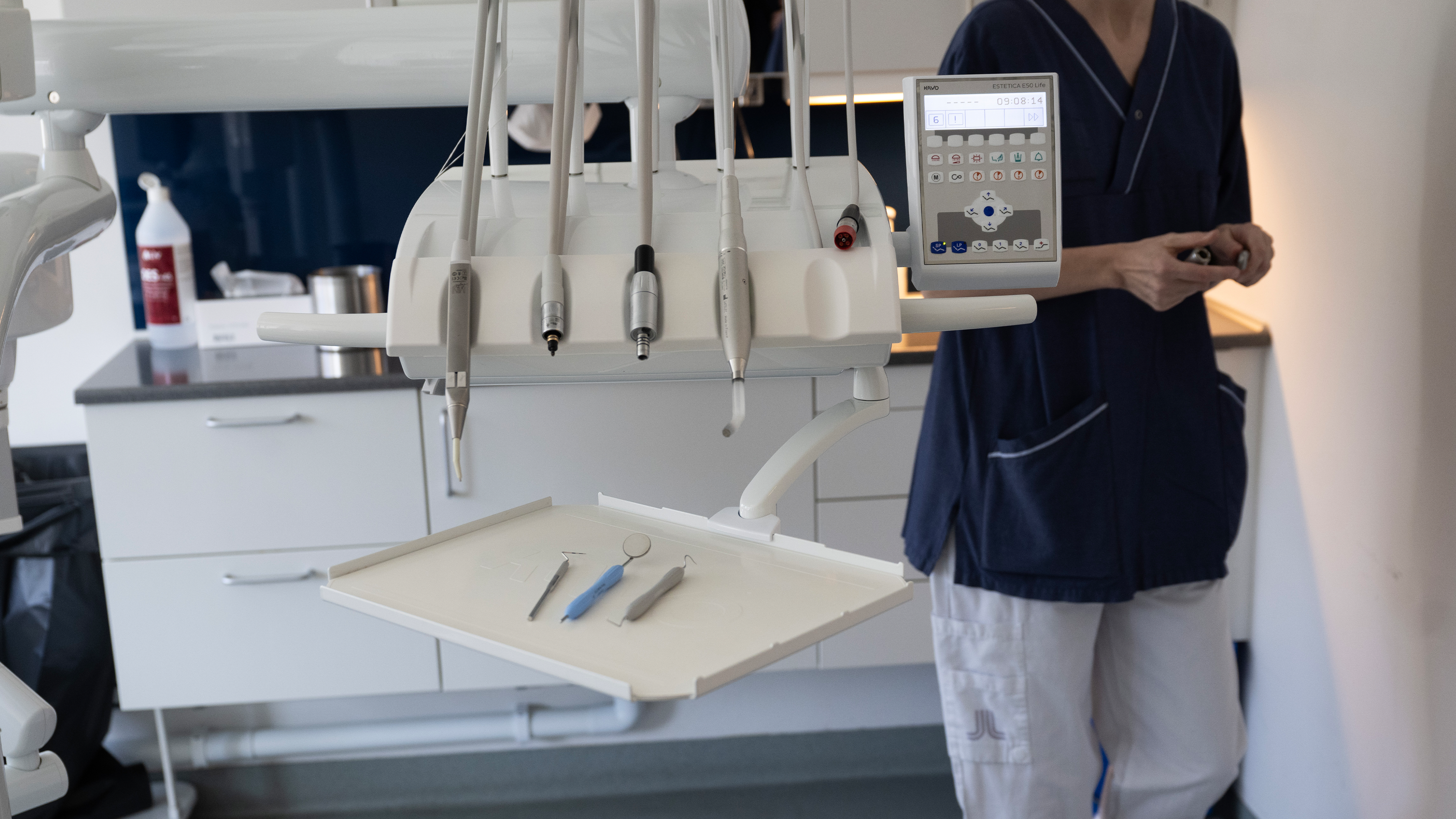 Tandläkarmottagning med tandvårdsutrustning i förgrunden och en person i arbetskläder står vid skåp i bakgrunden.