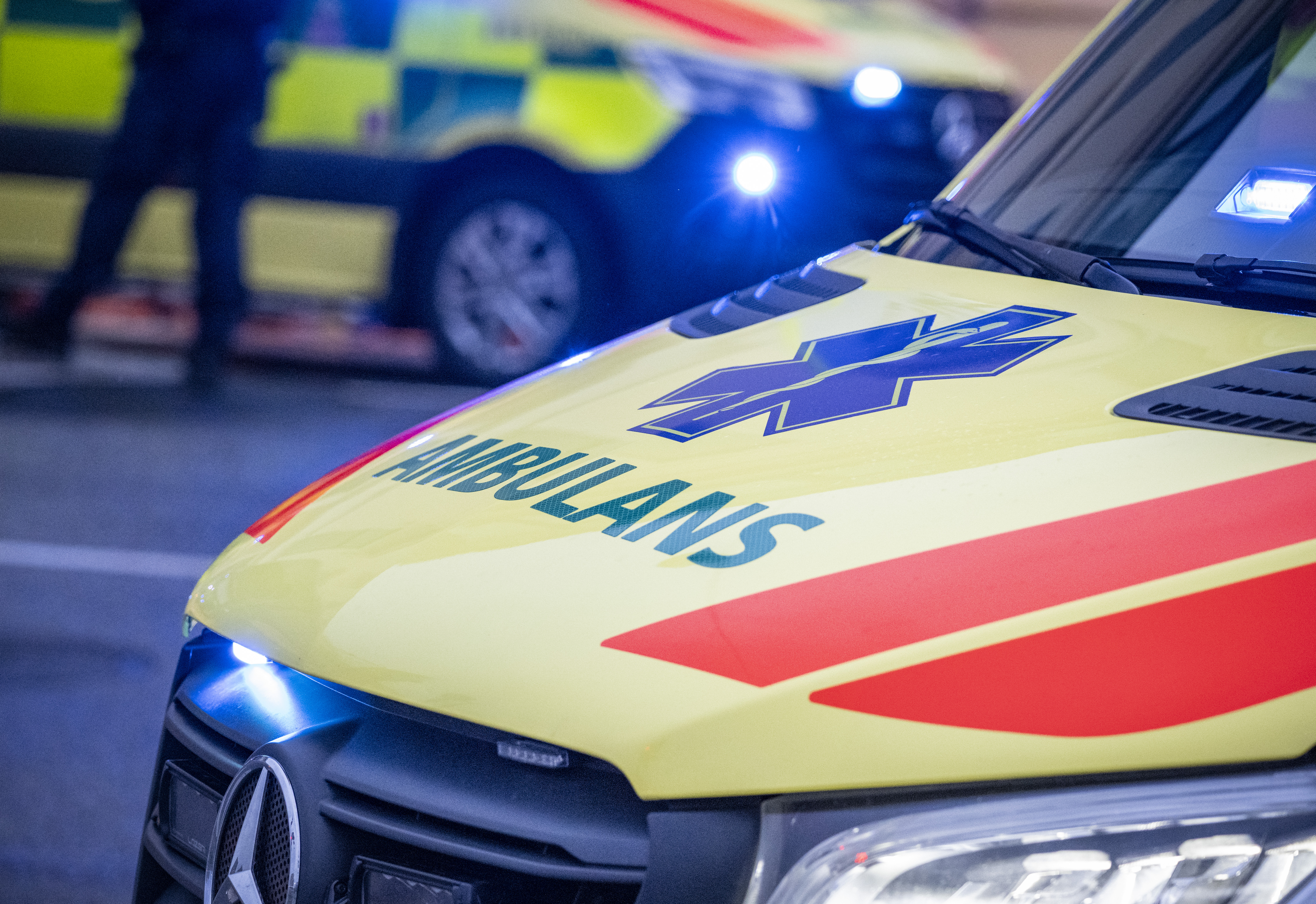 Ambulansens front med texten "AMBULANS" och blått vårdtecken, en annan utryckningsbil syns plötsligt i bakgrunden.