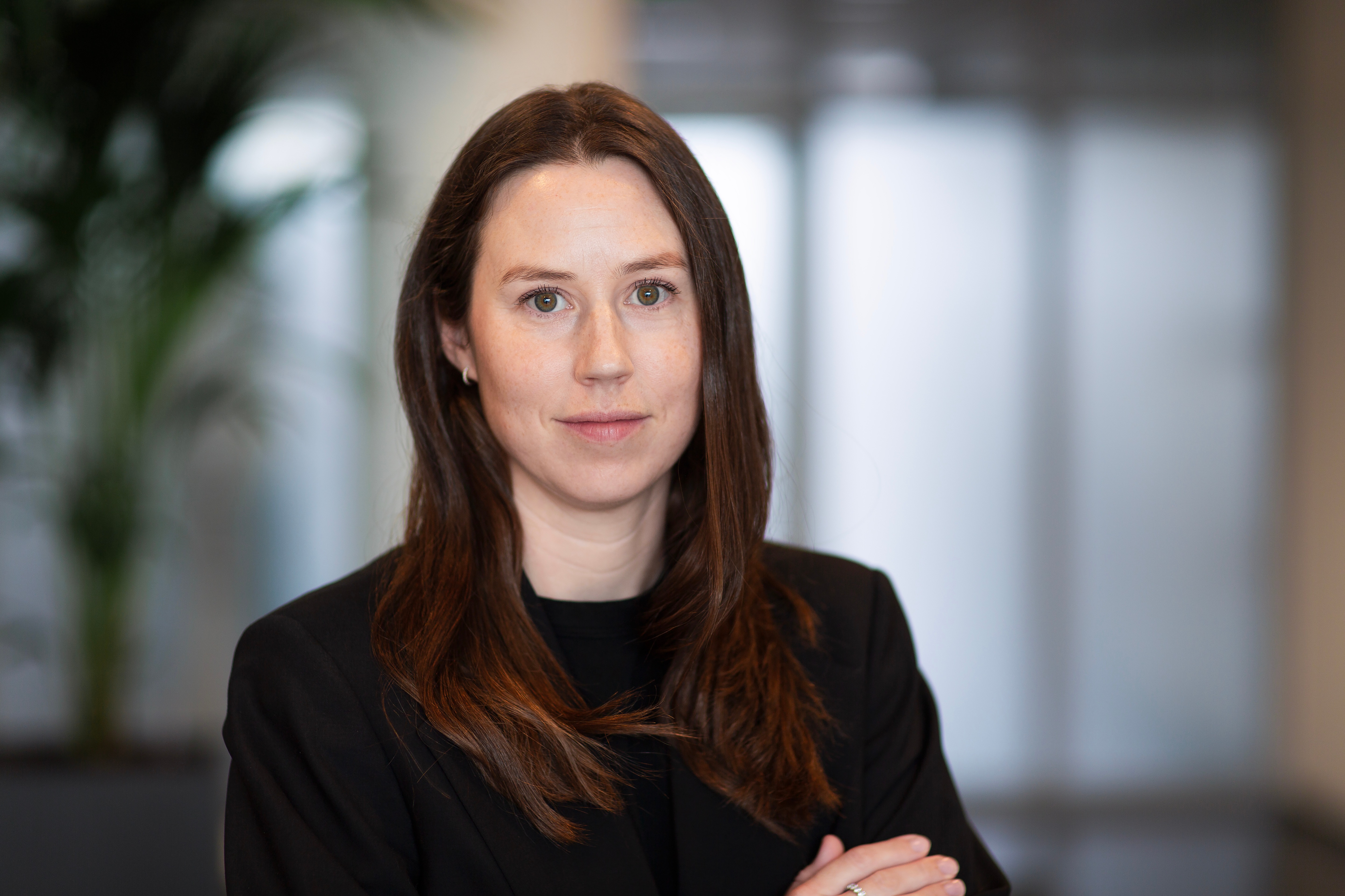 En kvinna med långt brunt hår och svart kavaj står inomhus med neutral min. Bakgrunden är suddig.