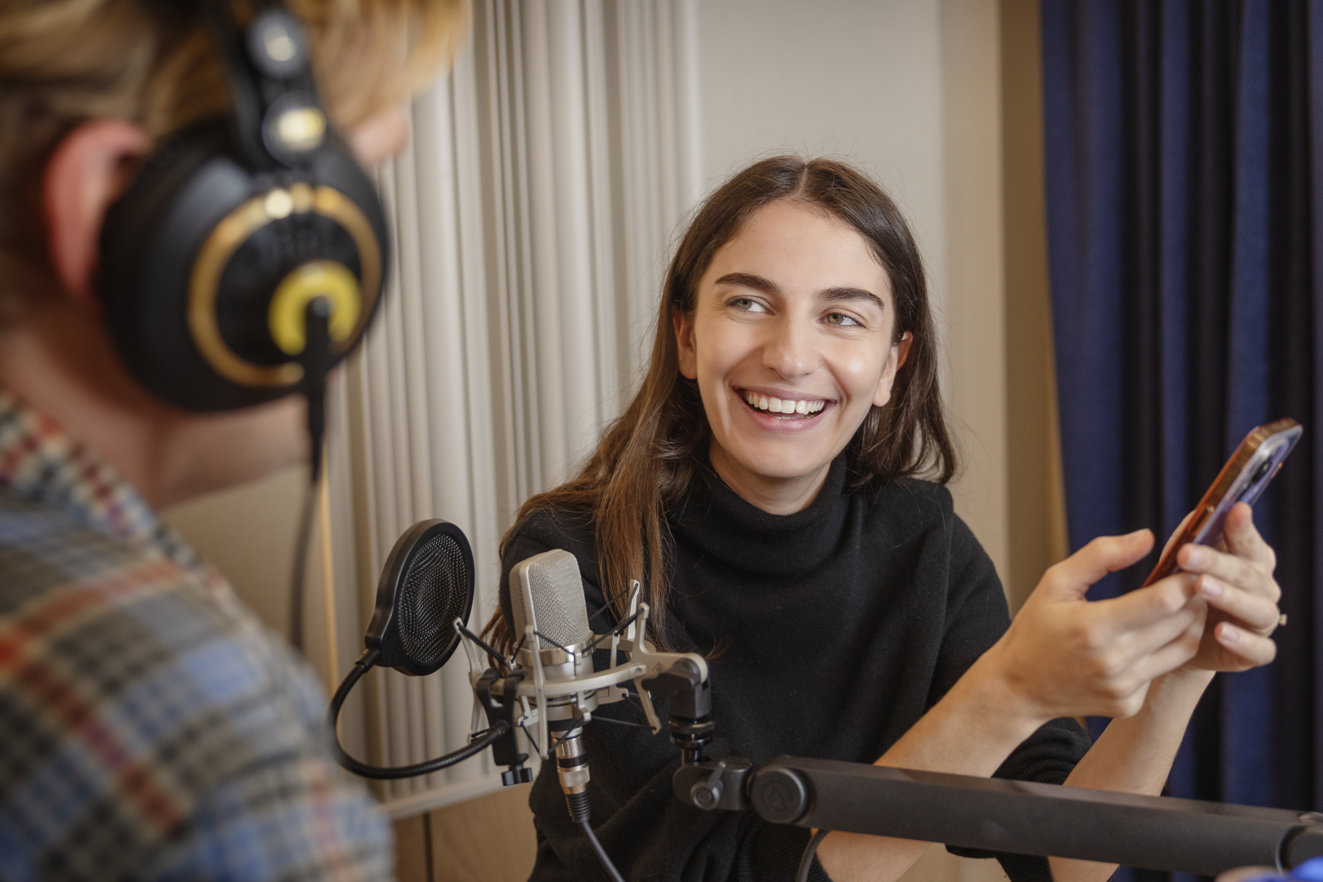 En kvinna håller en smartphone och ler mot en person med hörlurar vid ett bord med mikrofon och inspelningsutrustning.