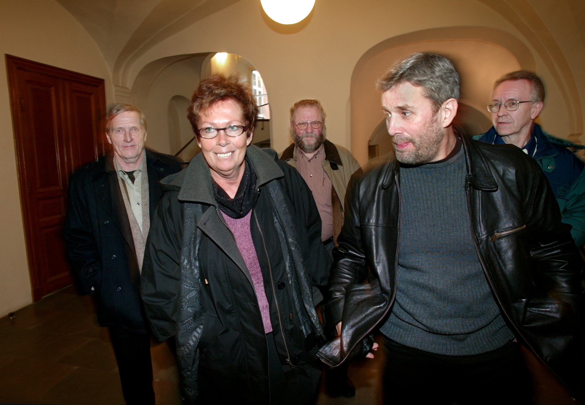 Fem vuxna med ytterkläder går tillsammans inomhus under valvade tak, en kvinna går först och ler, de andra följer efter.