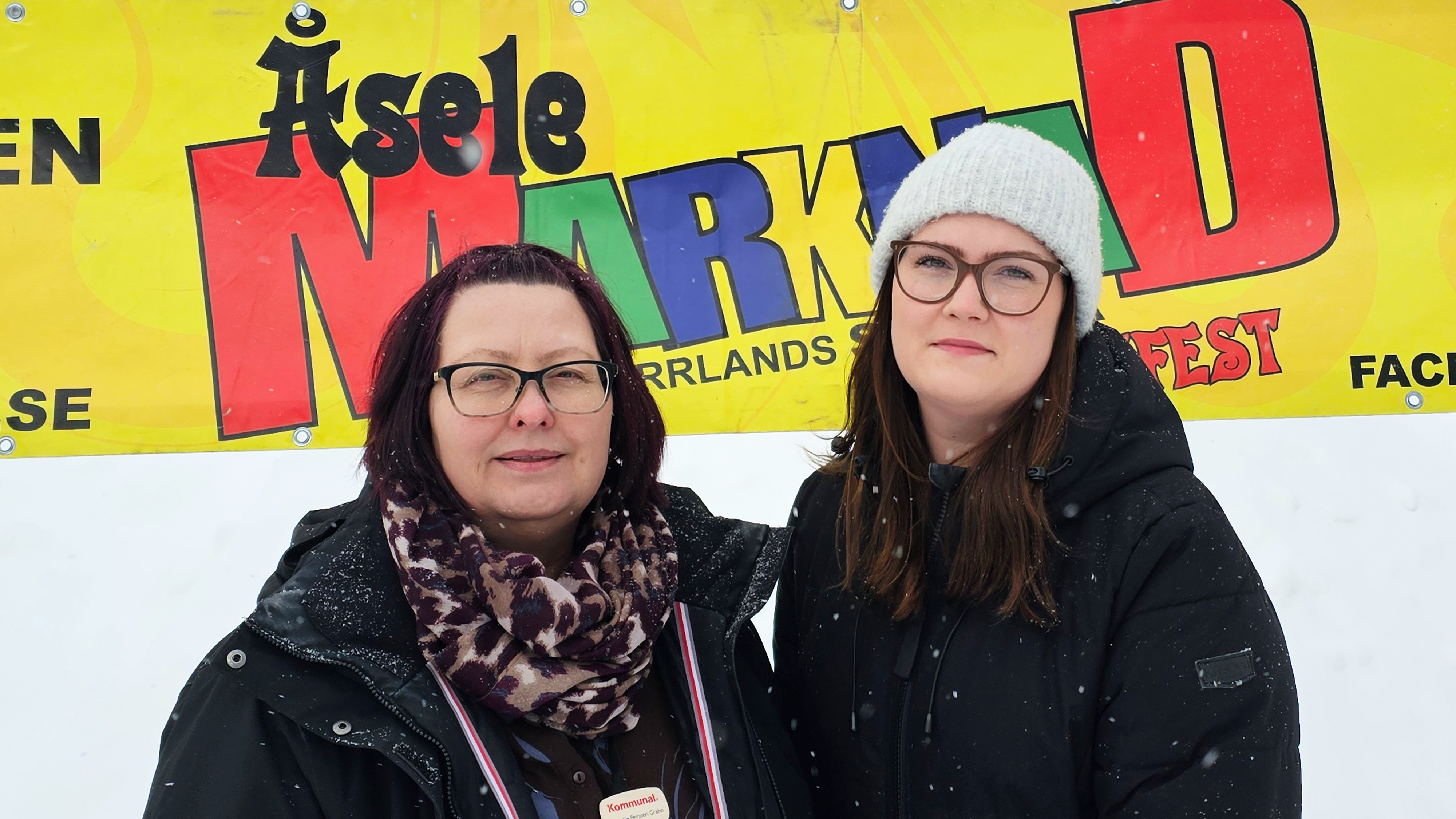 Två kvinnor står framför en gul skylt med texten "Åsele Marknad". Båda har vinterjacka, en har mössa och glasögon. Snö ute.