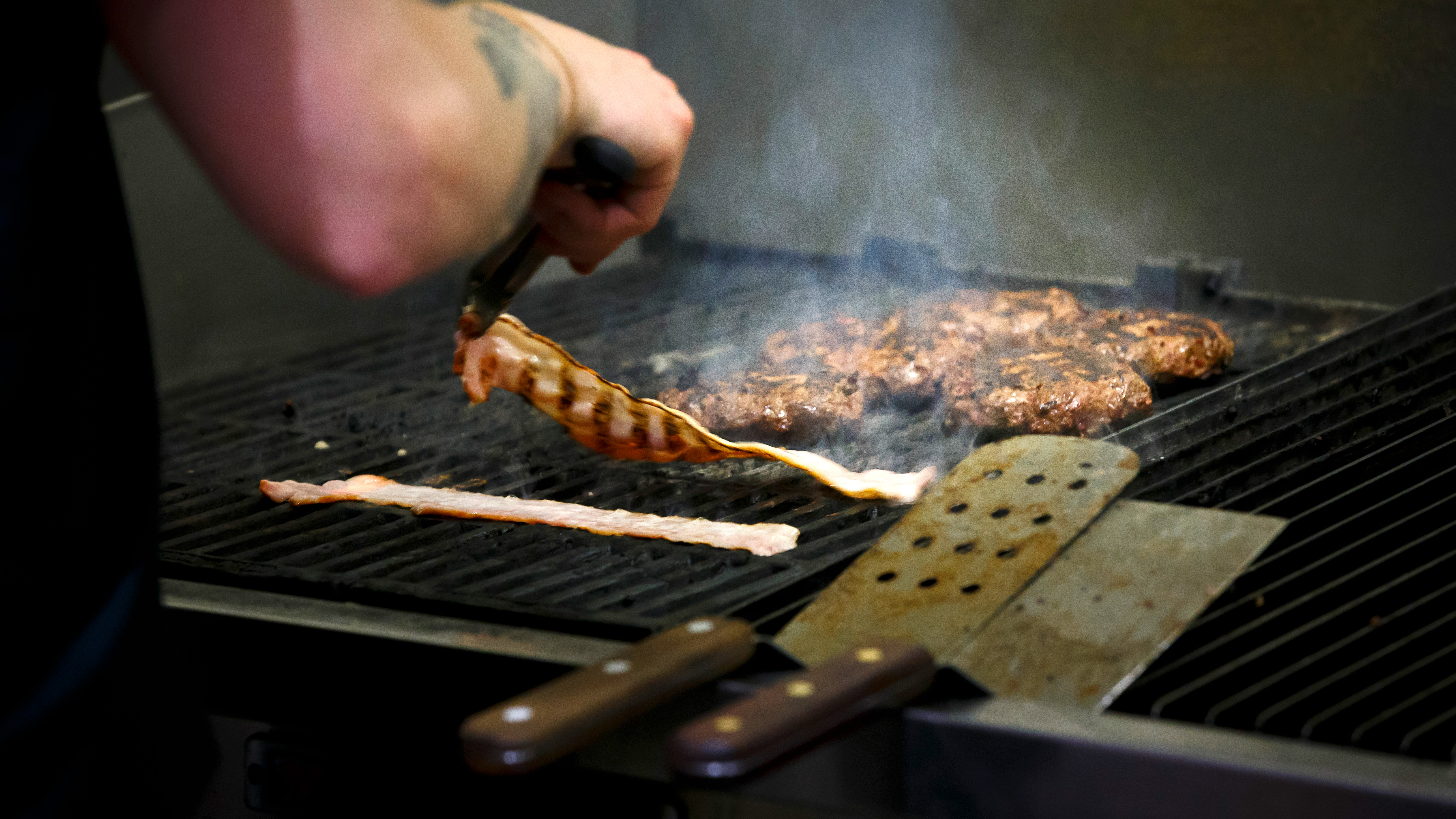En person använder en tång för att grilla bacon och hamburgare på en restauranggrill, med rök som stiger från maten.