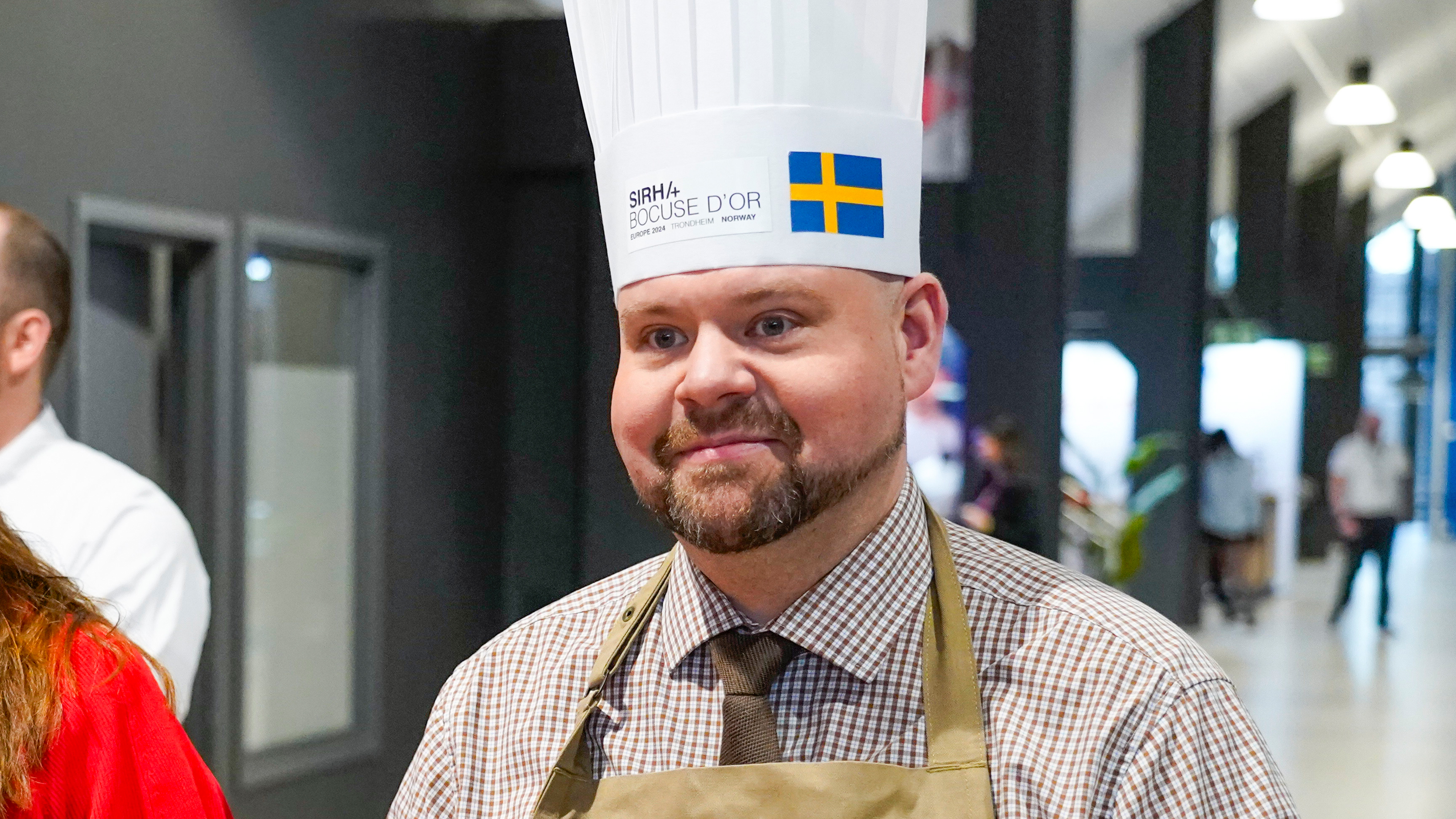 En man med kockmössa och förkläde står inomhus; mössan har en svensk flagga och texten "Sirha Bocuse d'Or".