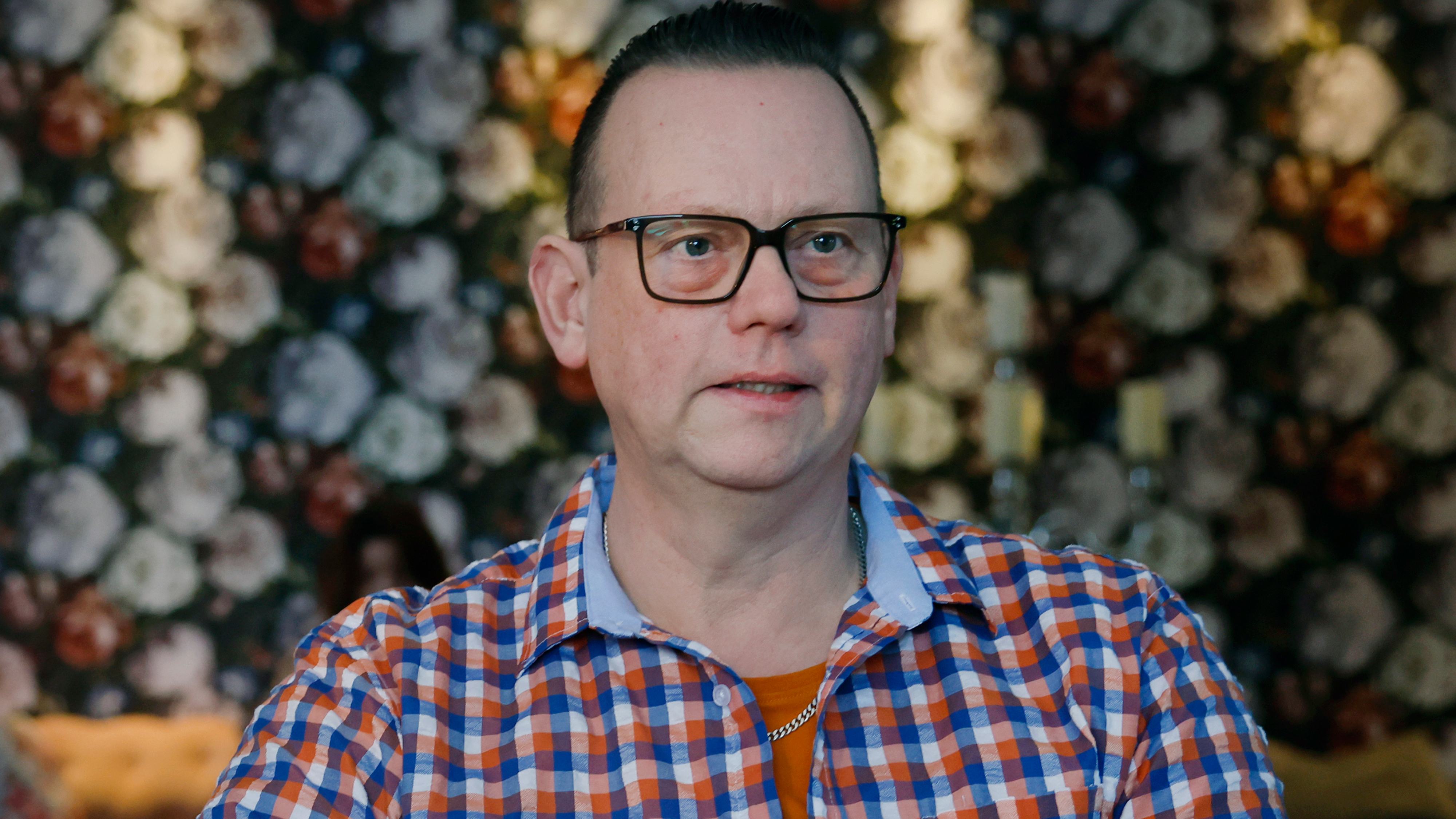 En person med glasögon och rutig skjorta sitter inomhus, med ett blommönster målat på baksidan.