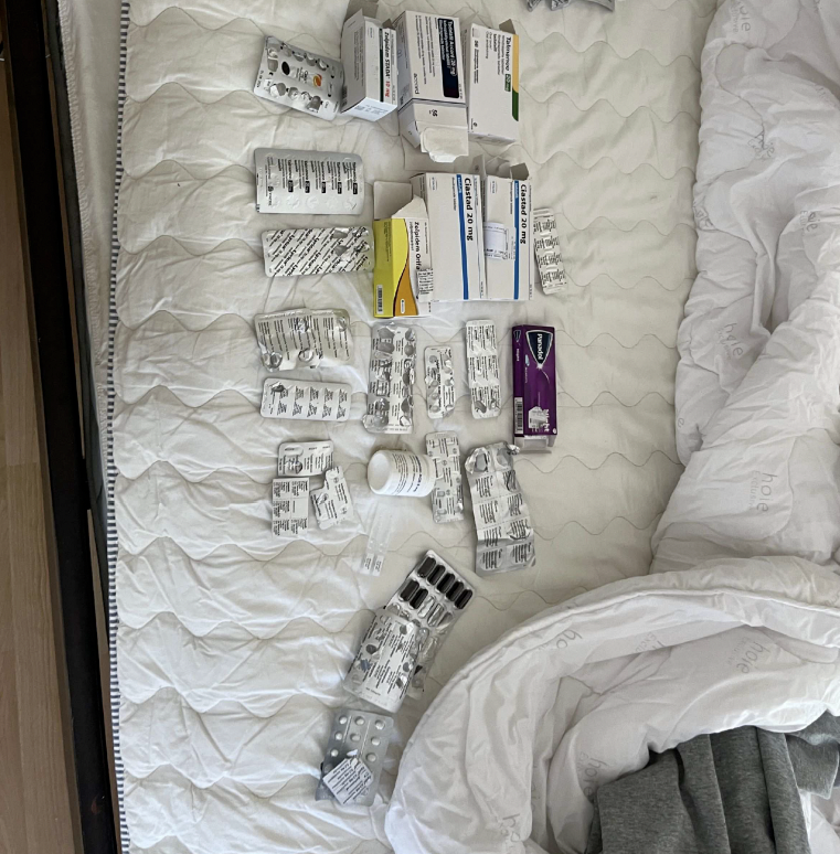 Flera askar och blisterförpackningar med medicin ligger på ett vitt täcke bredvid en obäddad säng med vit filt.