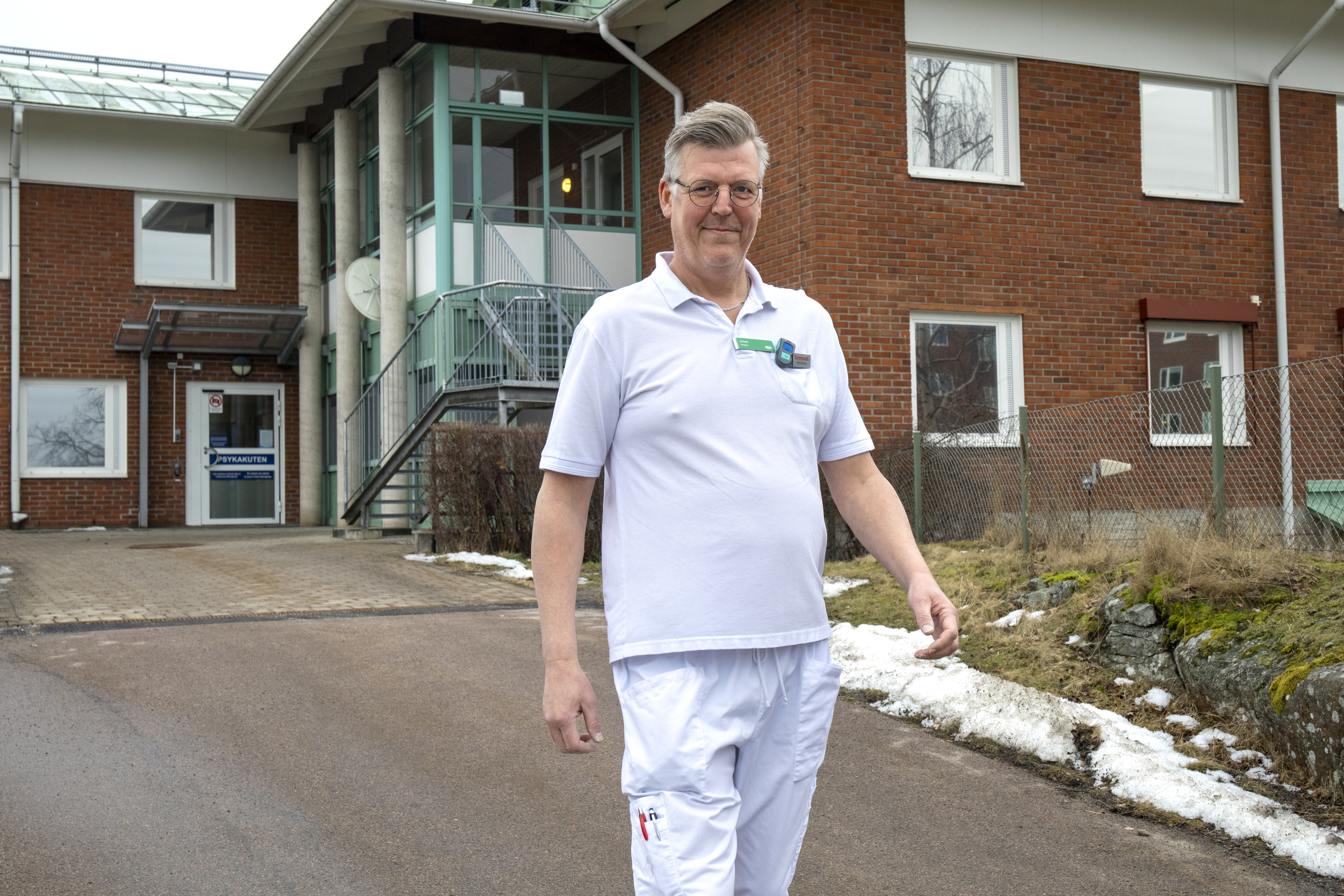 En man i vita medicinska kläder står utanför en byggnad av tegel med glasentré och snö på marken.