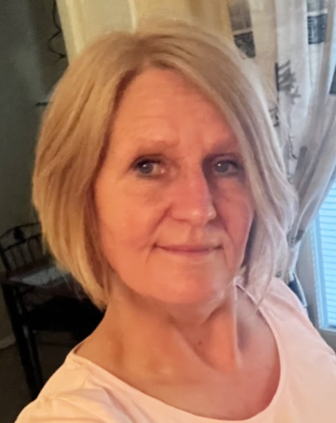 En kvinna med kort blont hår och ljusrosa tröja står inomhus och tittar mot kameran. Ett fönster med mönstrade gardiner syns bakom.