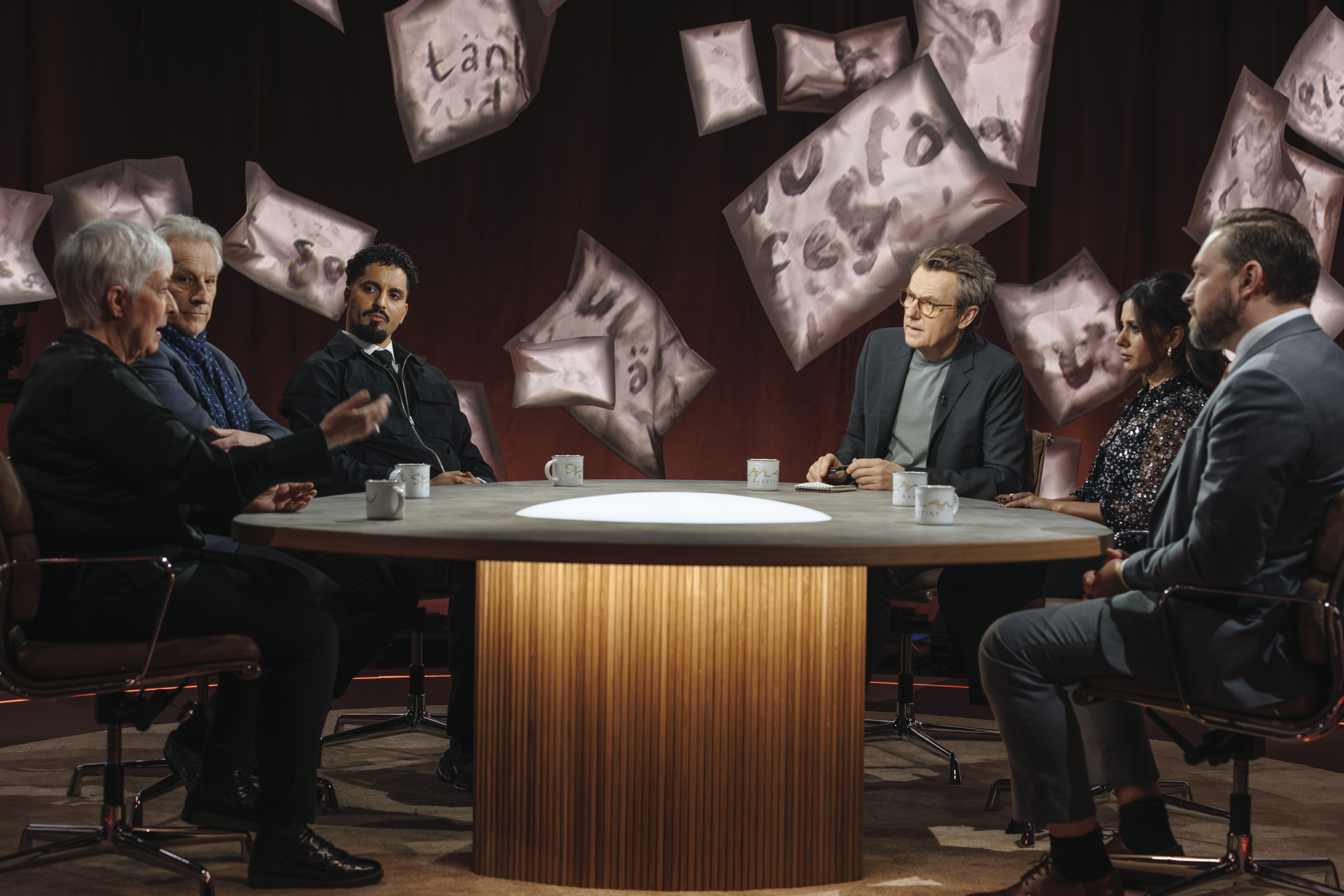 Sex personer sitter kring ett runt bord och pratar. Kaffekoppar står på bordet, konst hänger på väggen bakom dem.