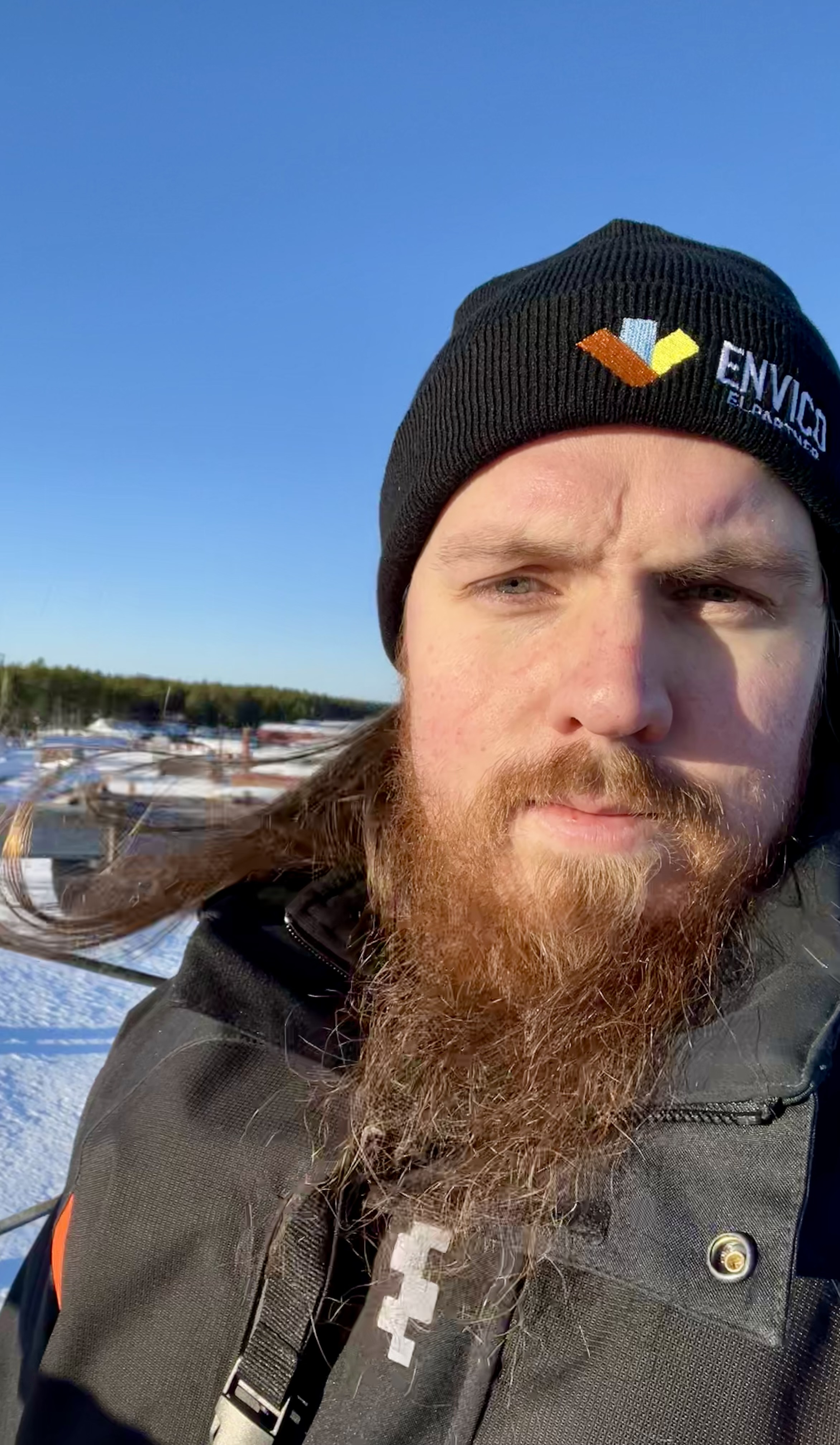 En skäggig person med svart mössa och jacka står utomhus en vinterdag med snö och hustak i bakgrunden.