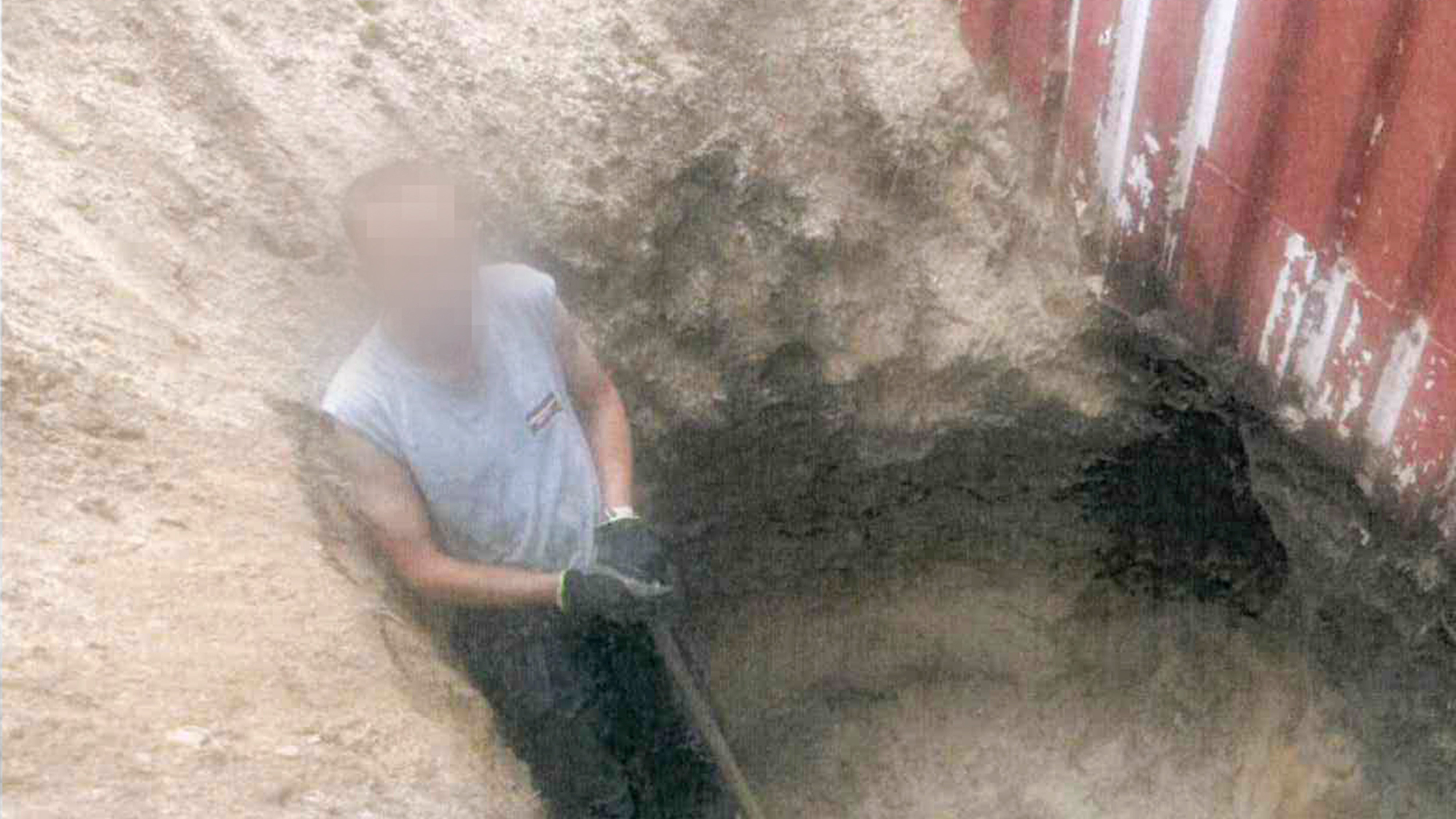 En person med suddigt ansikte gräver ett djupt hål i sandjord bredvid en delvis nedgrävd fraktcontainer.