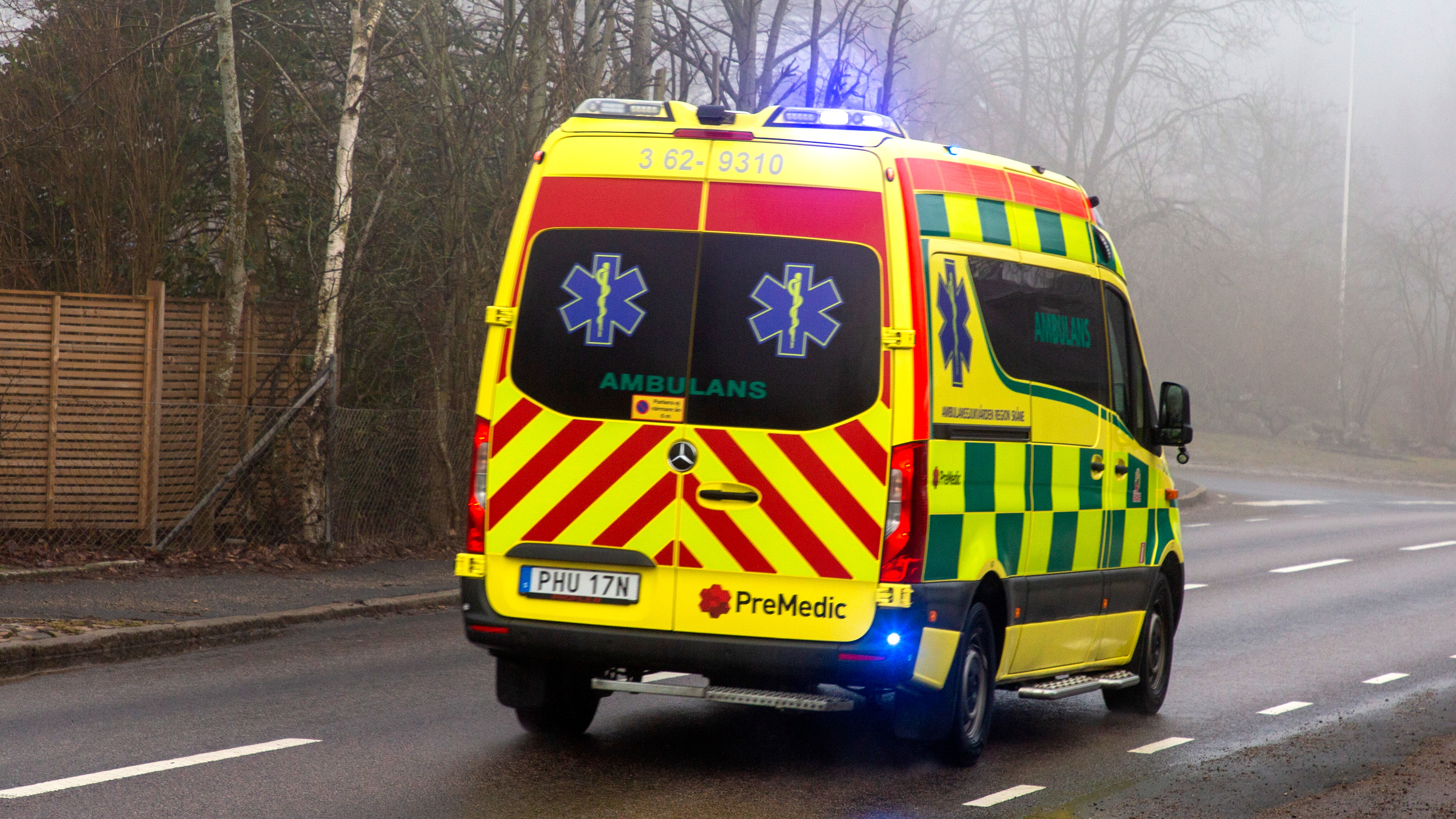 En gul och grön ambulans med blinkande ljus kör på en väg bredvid träd och ett staket.