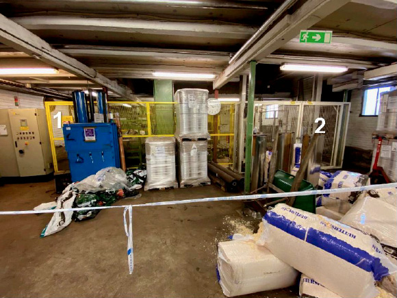 Industriellt lager med maskiner, staplade pallar i plast, kontrollpaneler och förpackningsmaterial på golvet; två sektioner märkta 1 och 2.