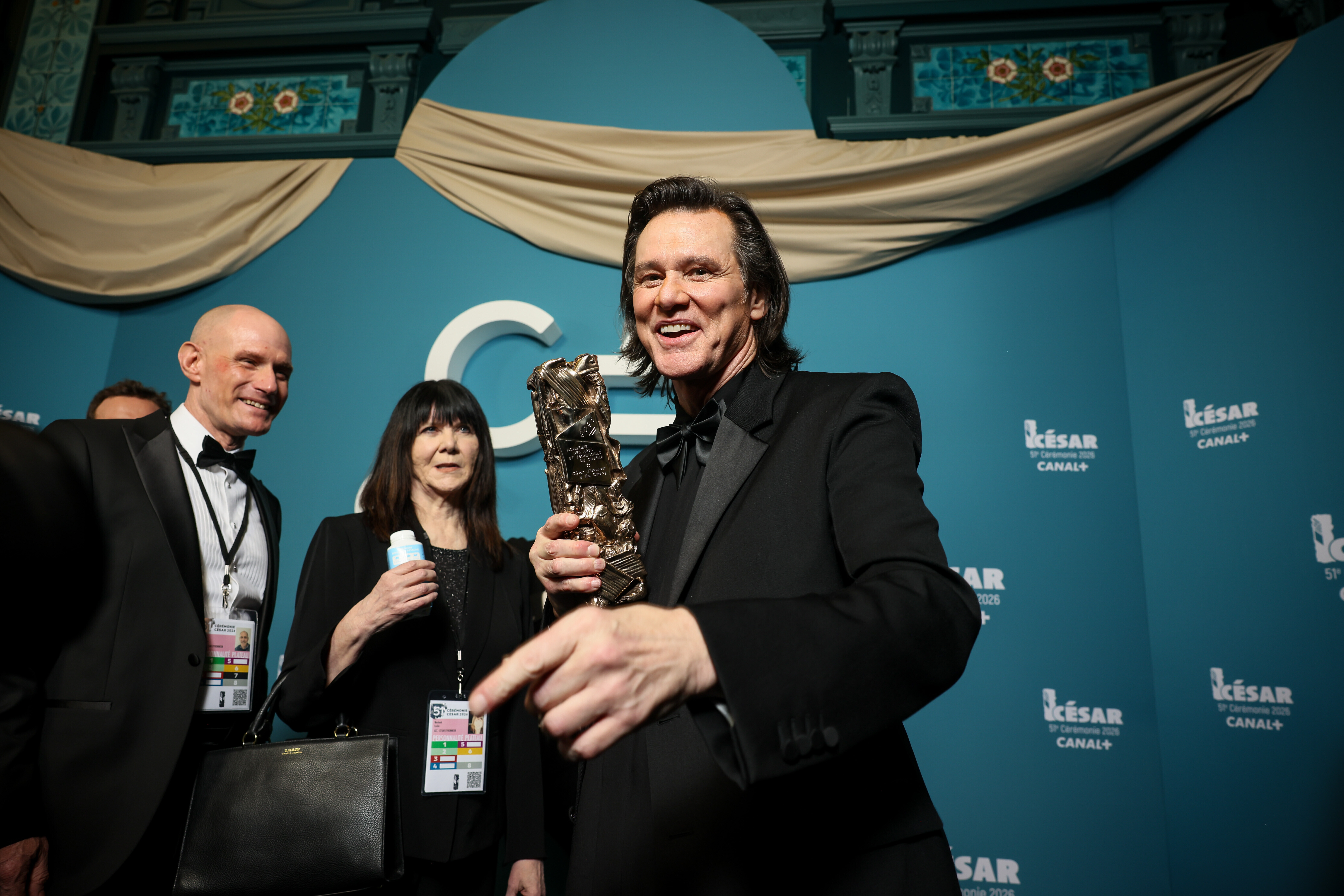 En man i svart kostym håller en Césarstatyett och ler mot kameran framför en blå bakgrund med två andra personer.