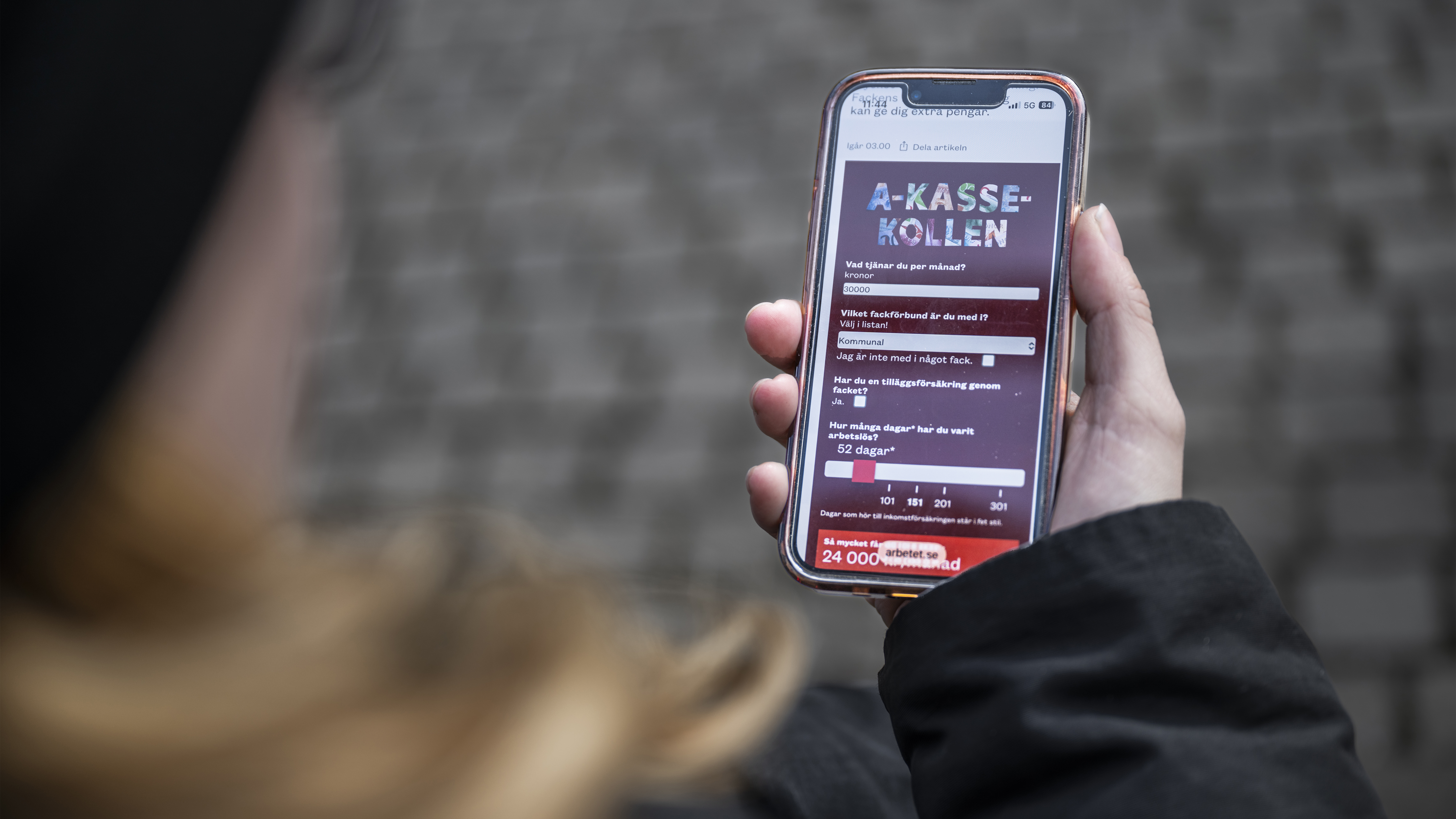 En person håller i en smartphone som visar webbplatsen ”A-kassekollen” med ett frågeformulär på skärmen.