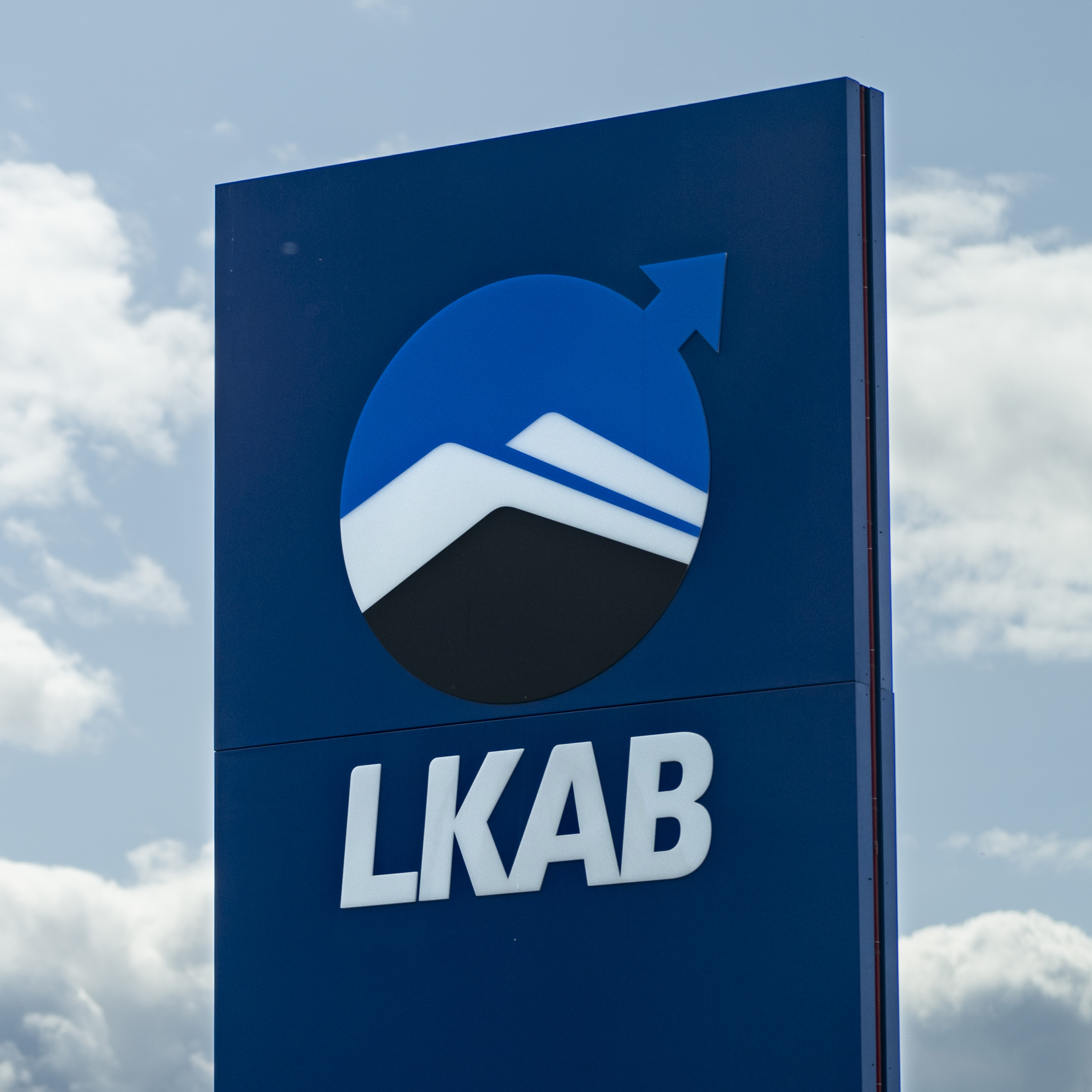 En blå skylt med LKAB-logotypen, stiliserade berg och en uppåtriktad pil, mot en himmel med några moln.