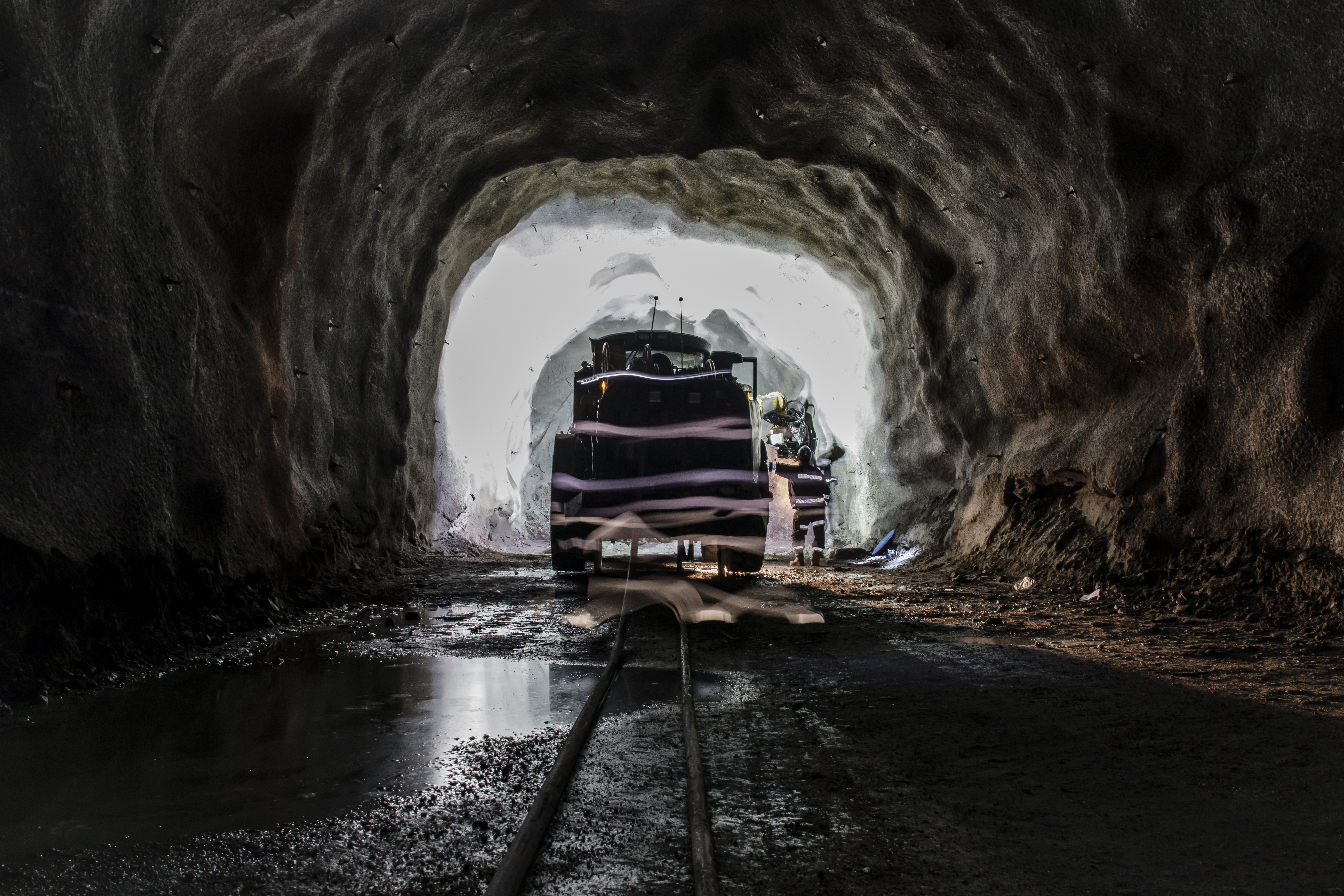 En gruvtunnel med bilar på spåren och två arbetare som står vid ingången till tunneln, upplysta av ljus utifrån.