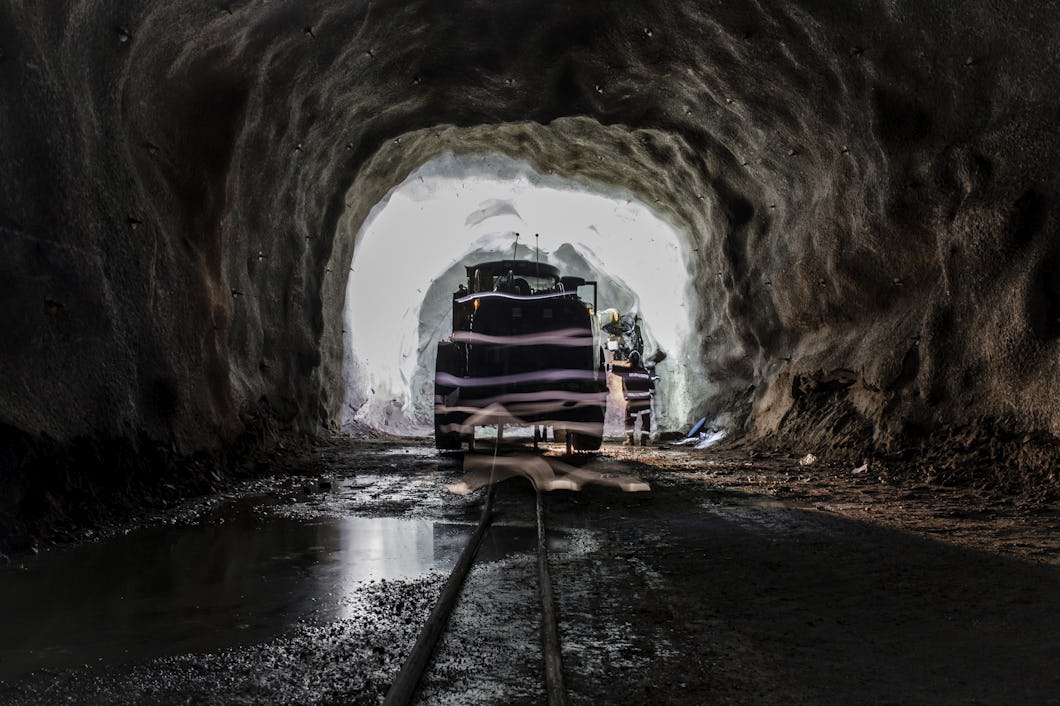 En gruvtunnel med bilar på spåren och två arbetare som står vid ingången till tunneln, upplysta av ljus utifrån.