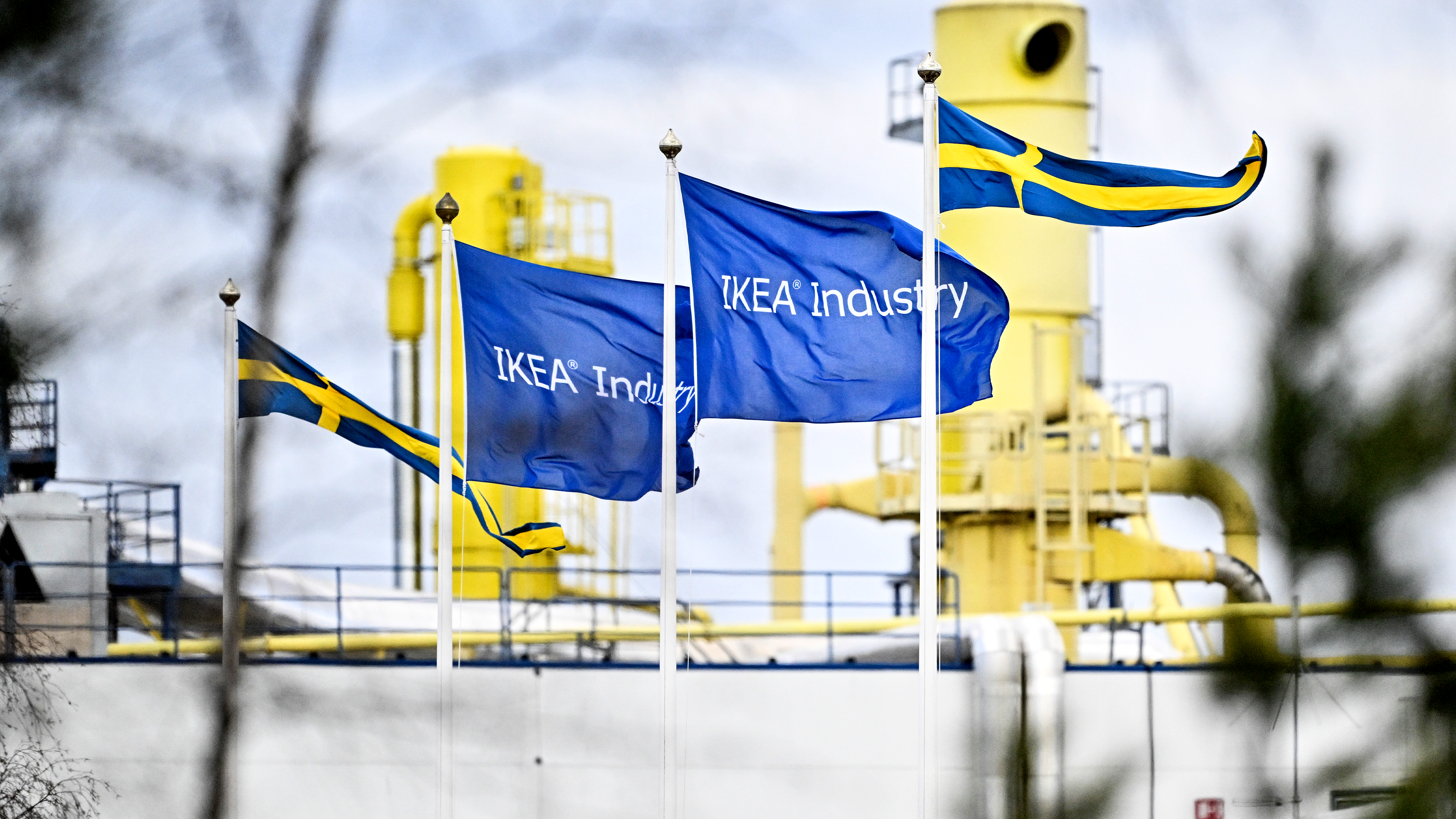 Tre IKEA Industry-flaggor och två svenska flaggor vajar framför en gul industribyggnad.