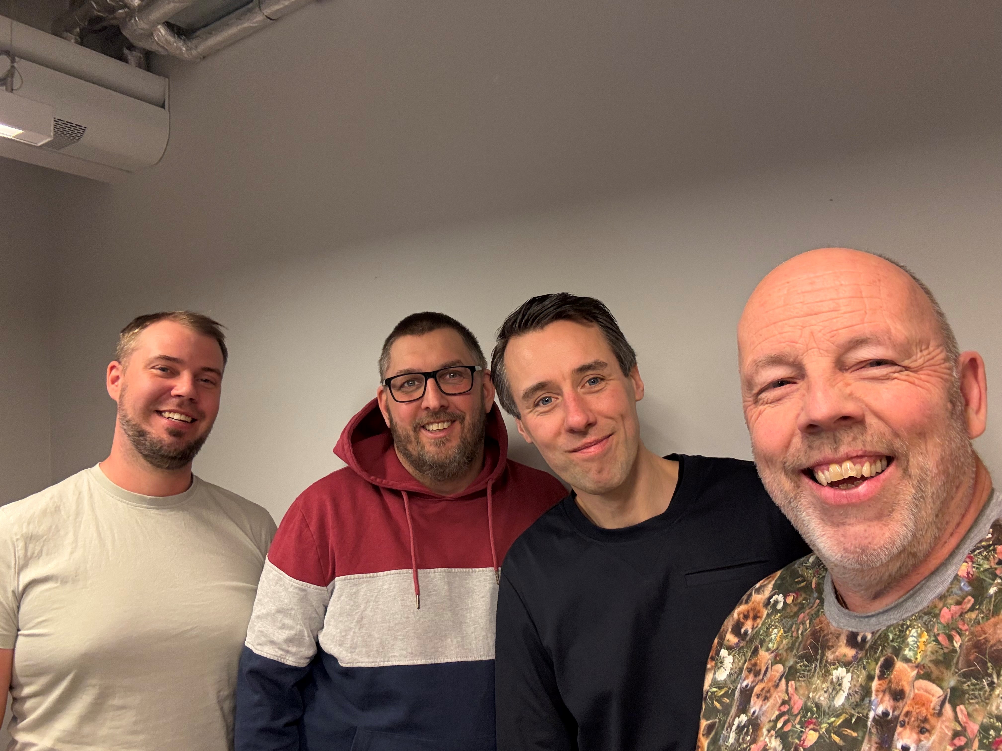 Fyra män står inomhus framför en ljusgrå vägg och ser mot kameran.