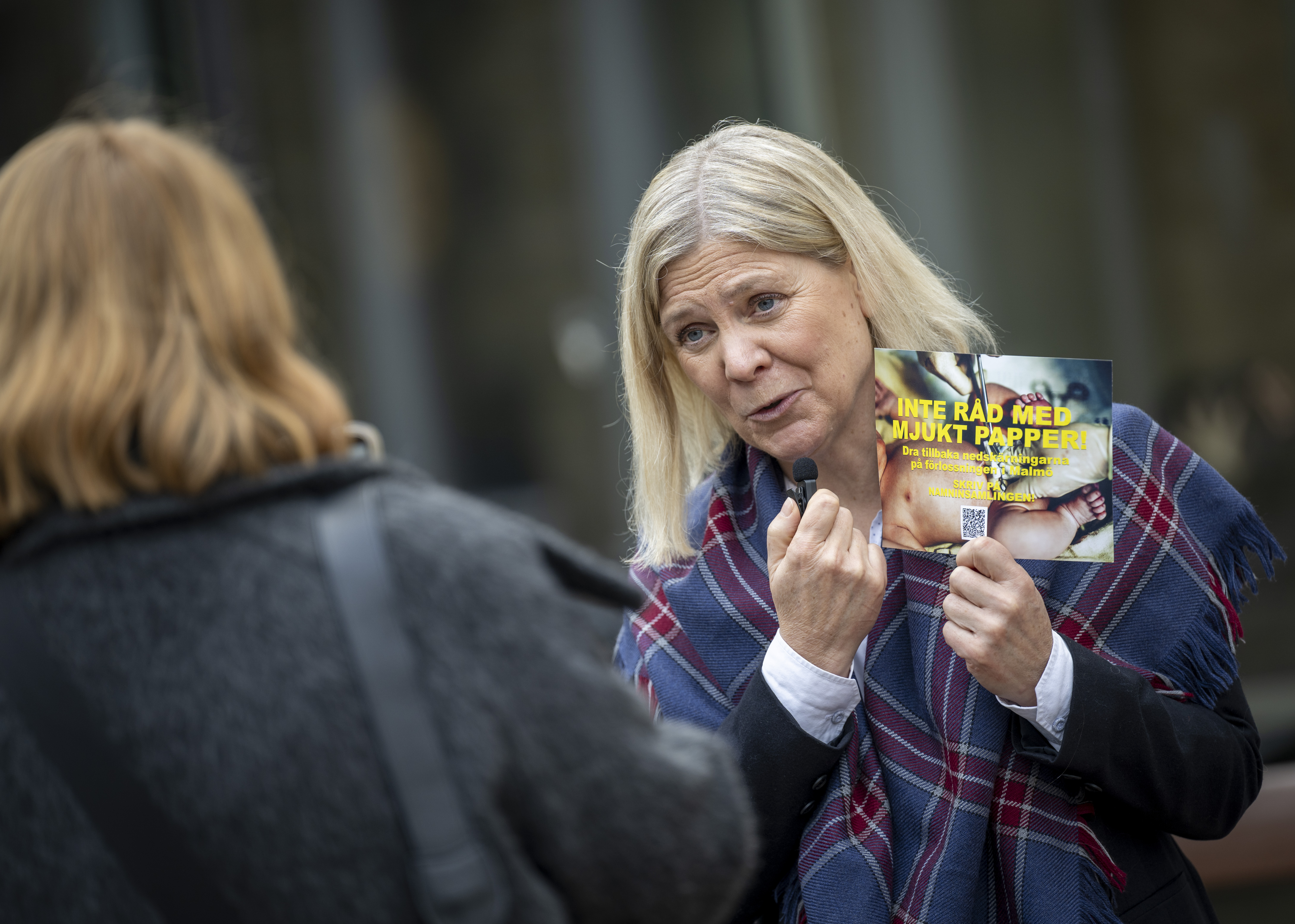 En kvinna håller i ett flygblad och pratar med en annan person utomhus. Flygbladet har en bild och gul text på svenska.