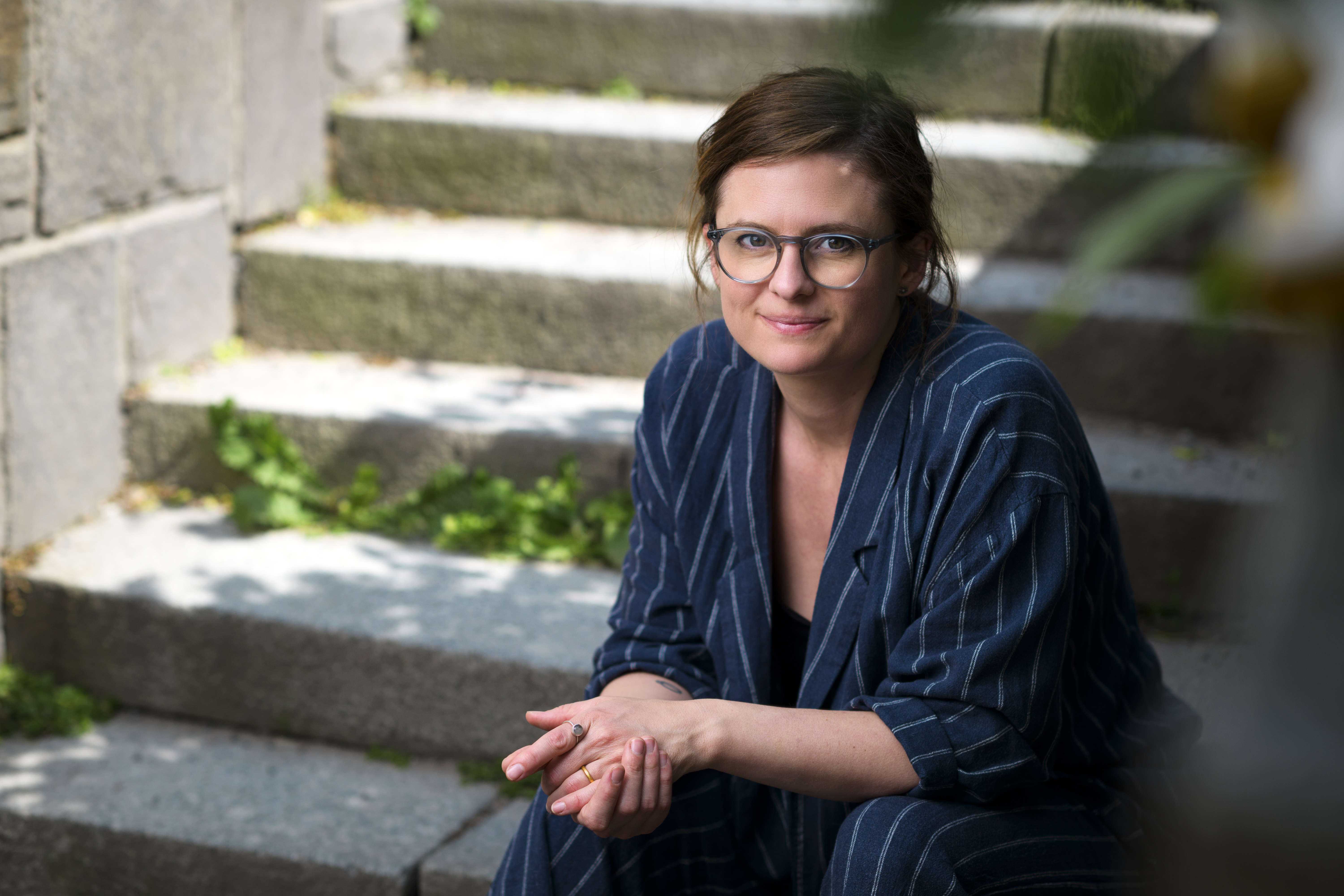 Författaren Johanna Holmström Svedén med glasögon och brunt hår sitter på utomhustrappor, har på sig en blå- och vitrandig outfit och tittar mot kameran.
