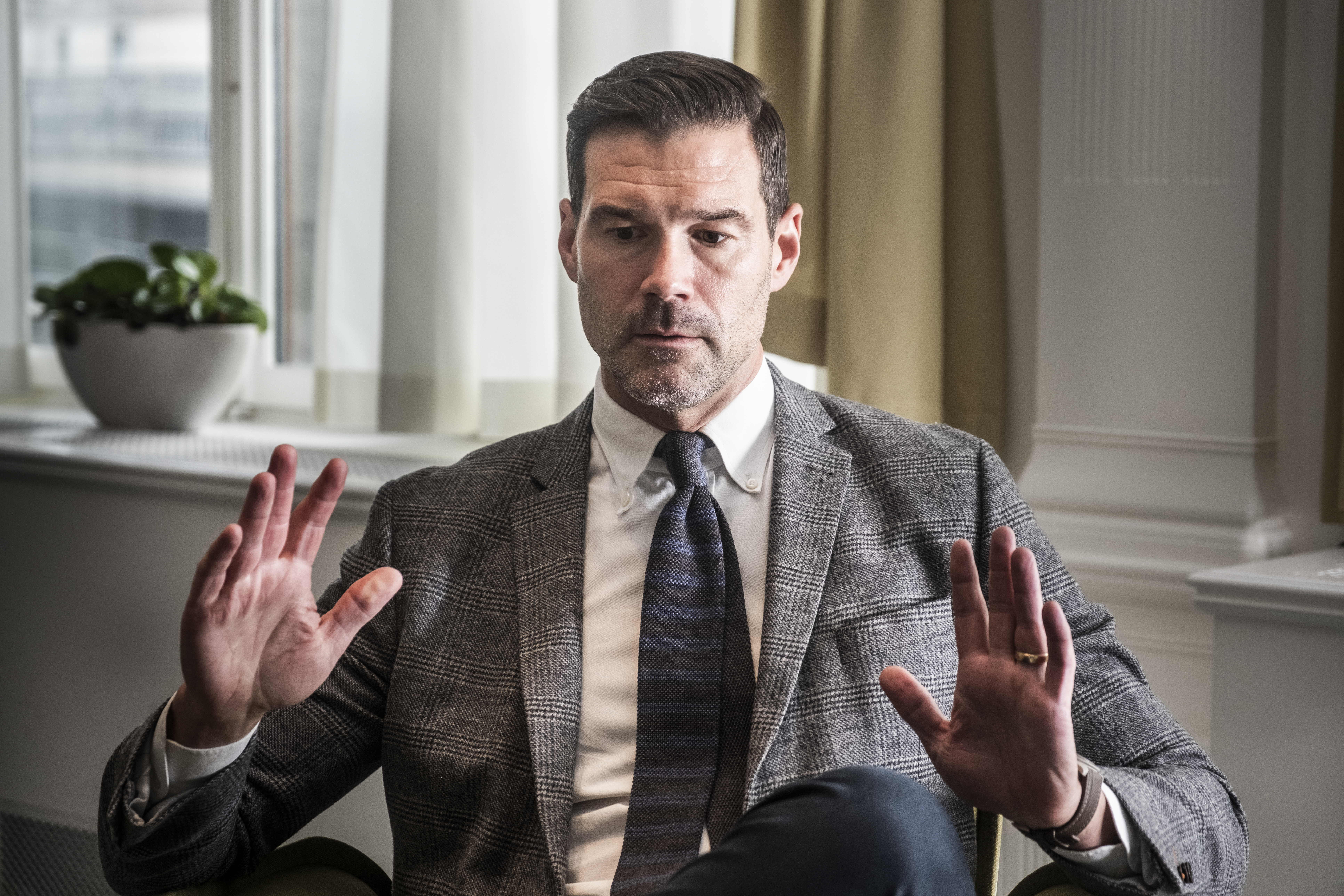 En man i grå kostym och randig slips sitter inomhus med händerna upphöjda. Ett fönster och en krukväxt syns bakom.