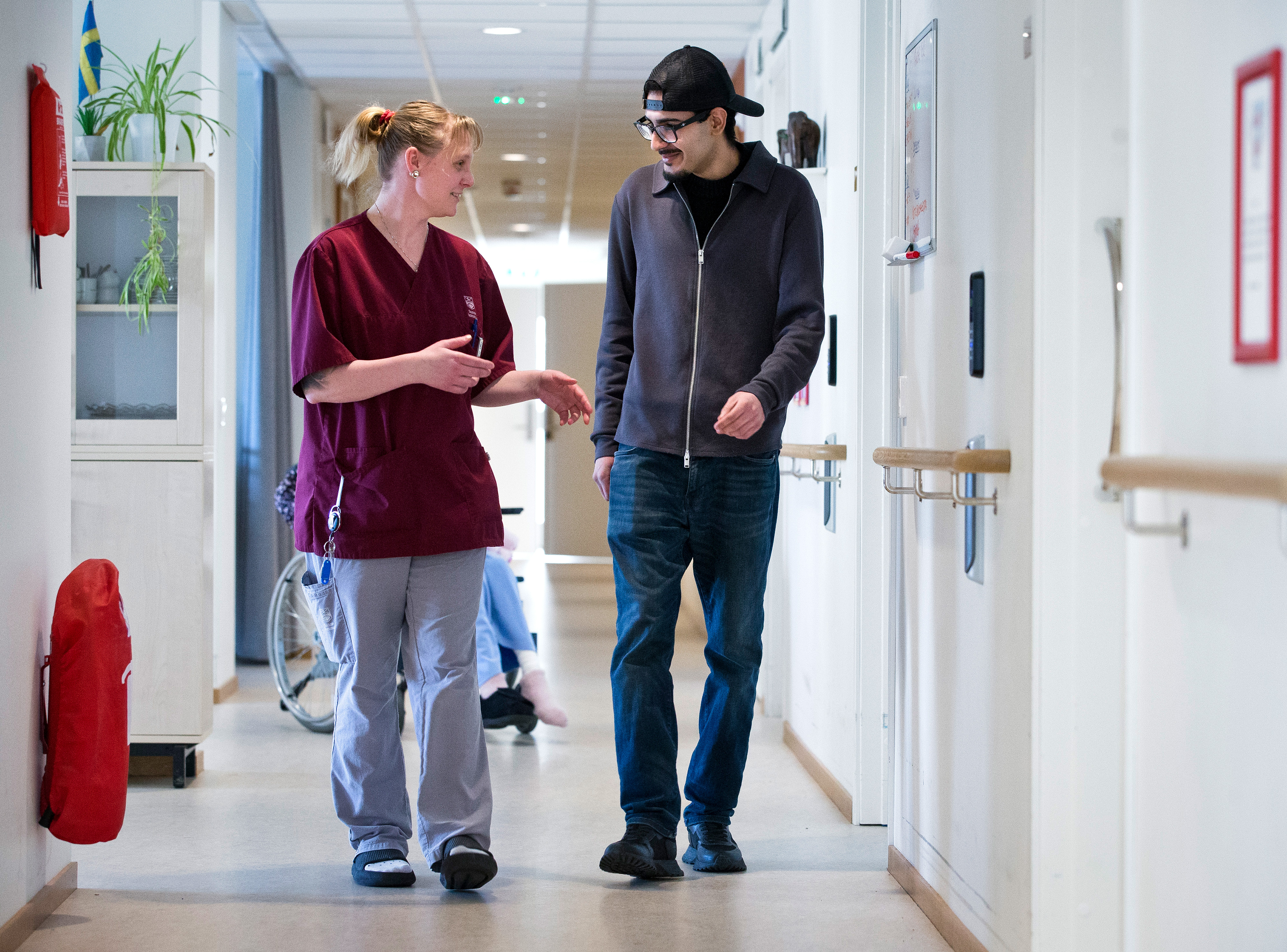 Angelica vårdarbetare i vinröda arbetskläder går och pratar med en Muhammed i lediga klädsel i en korridor, med rullstol och ytterligare en person i bakgrunden.