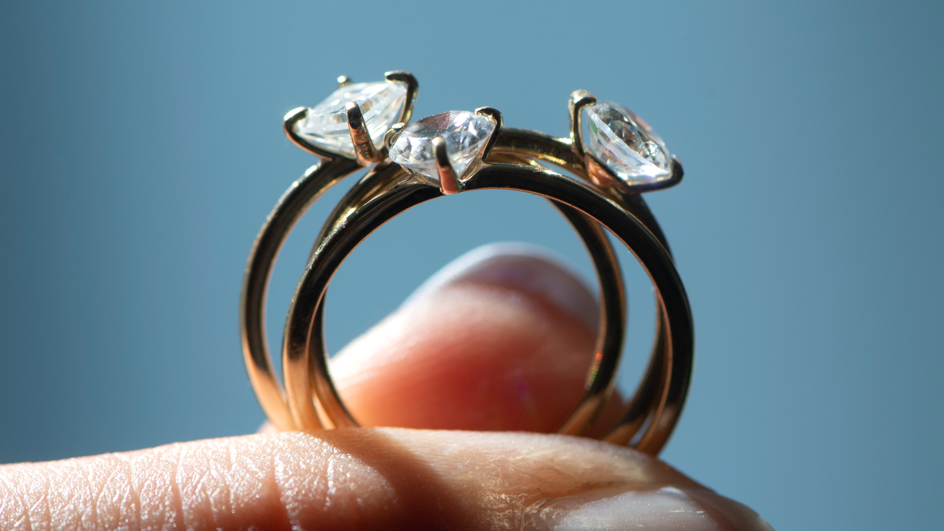 Närbild av två guldringar med stora, genomskinliga stenar som hålls mellan en person fingrar mot blå bakgrund.