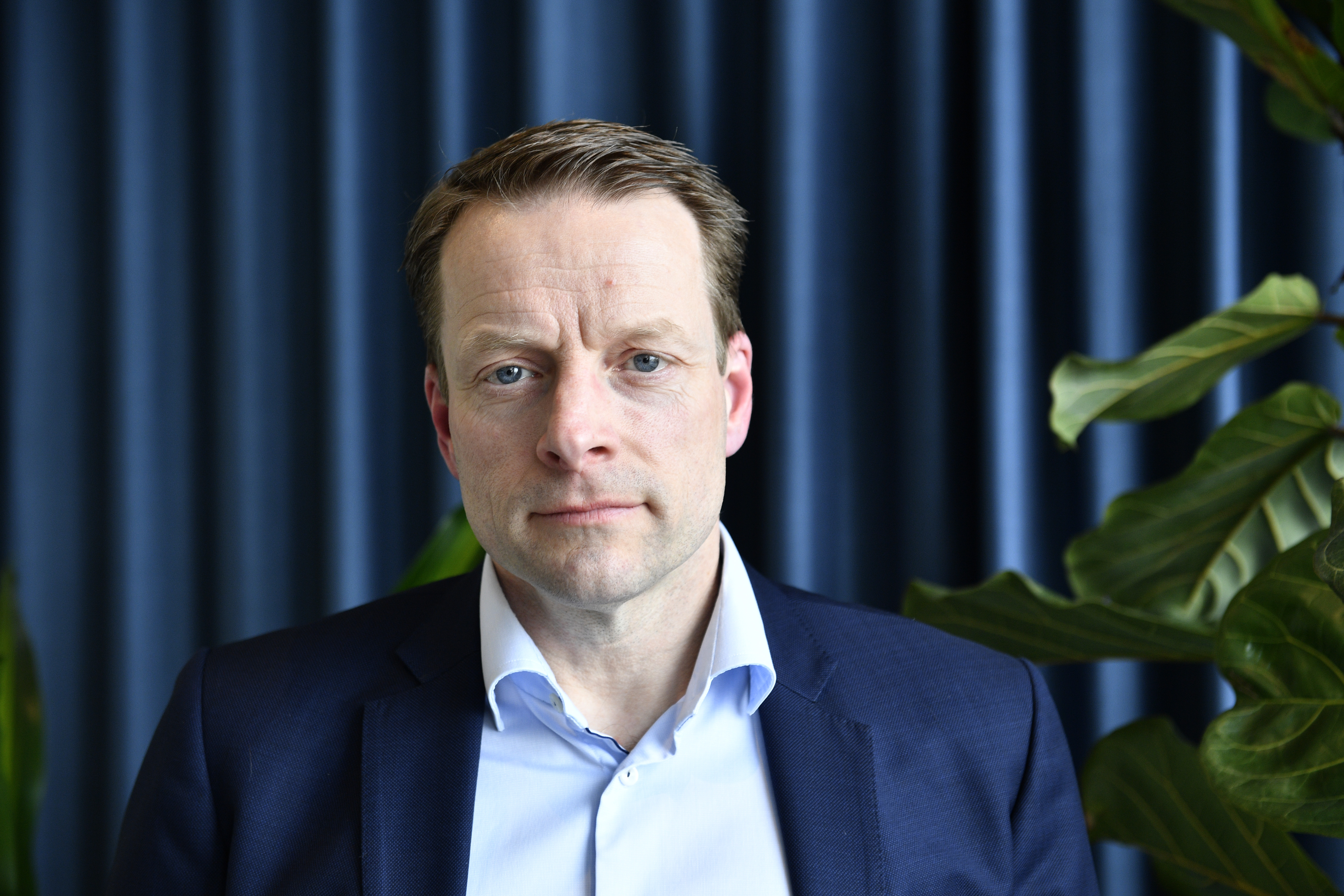 En man i mörk kostym och ljusblå skjorta står framför blå gardiner flankerade av gröna växter.