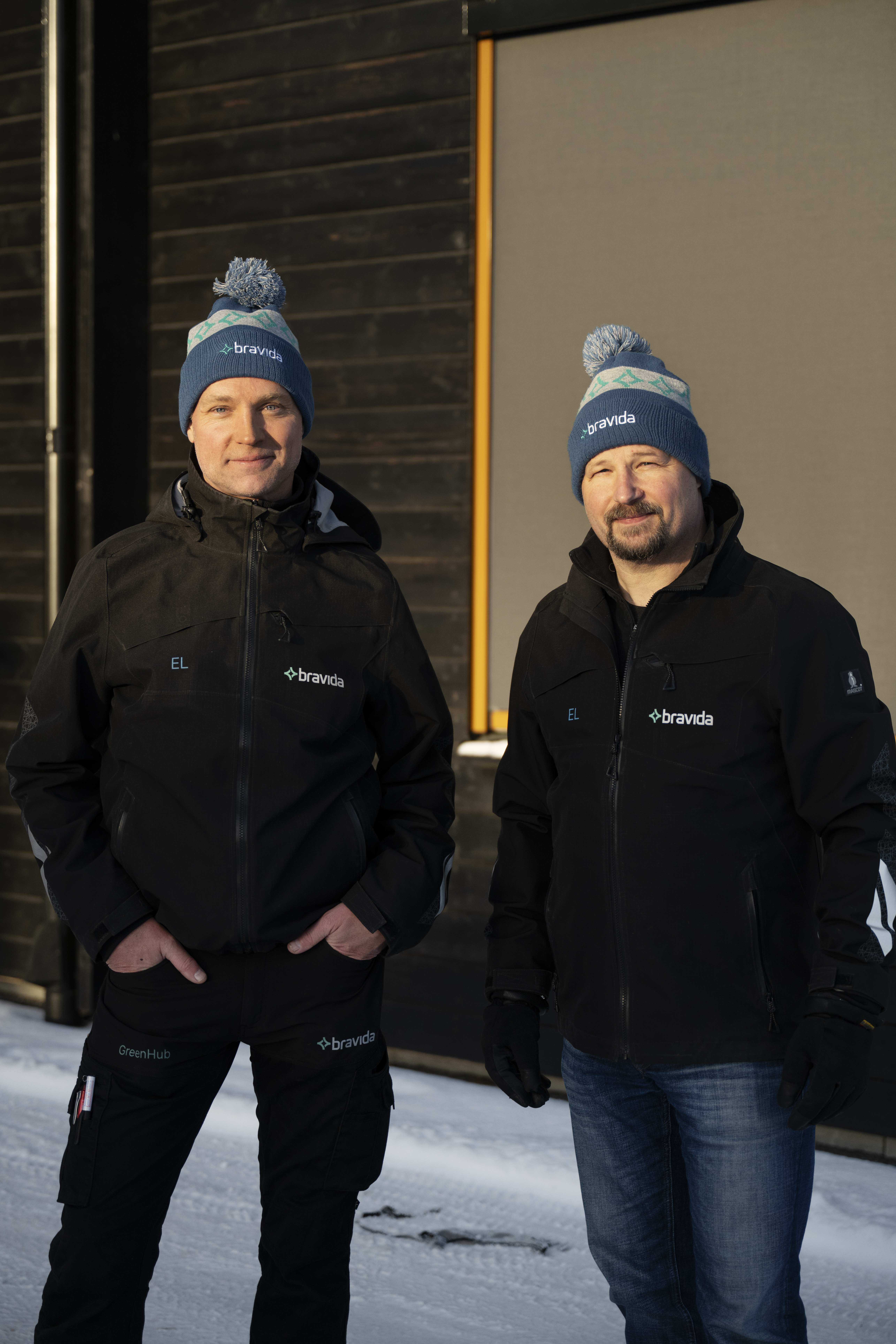 Två män med blå Bravida-mössor och svarta Bravida-jackor står utomhus i snö framför en mörk byggnad.
