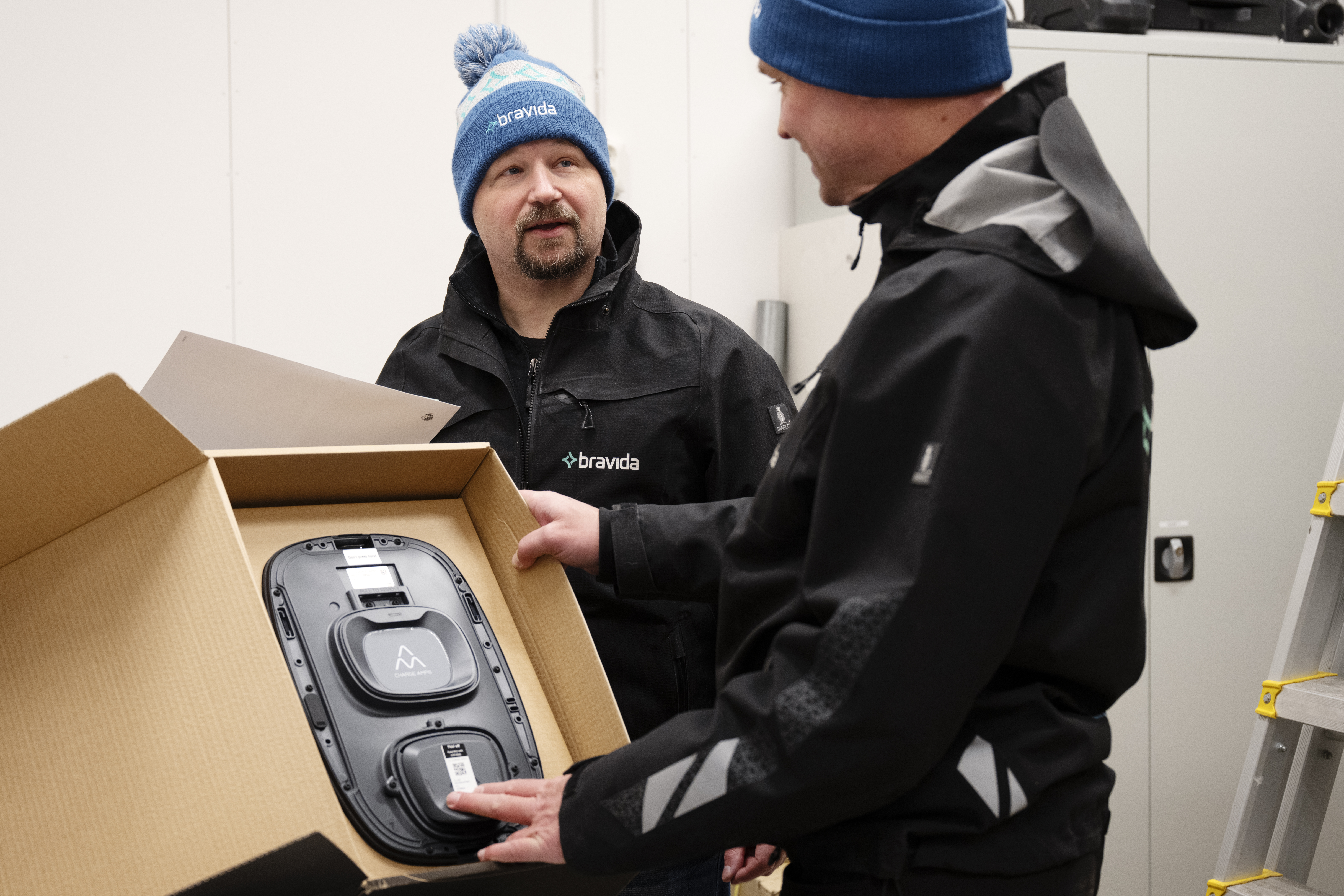 Två personer i jackor med logotyp och blå kepsar undersöker en svart elektronisk utrustning i en kartong inomhus.