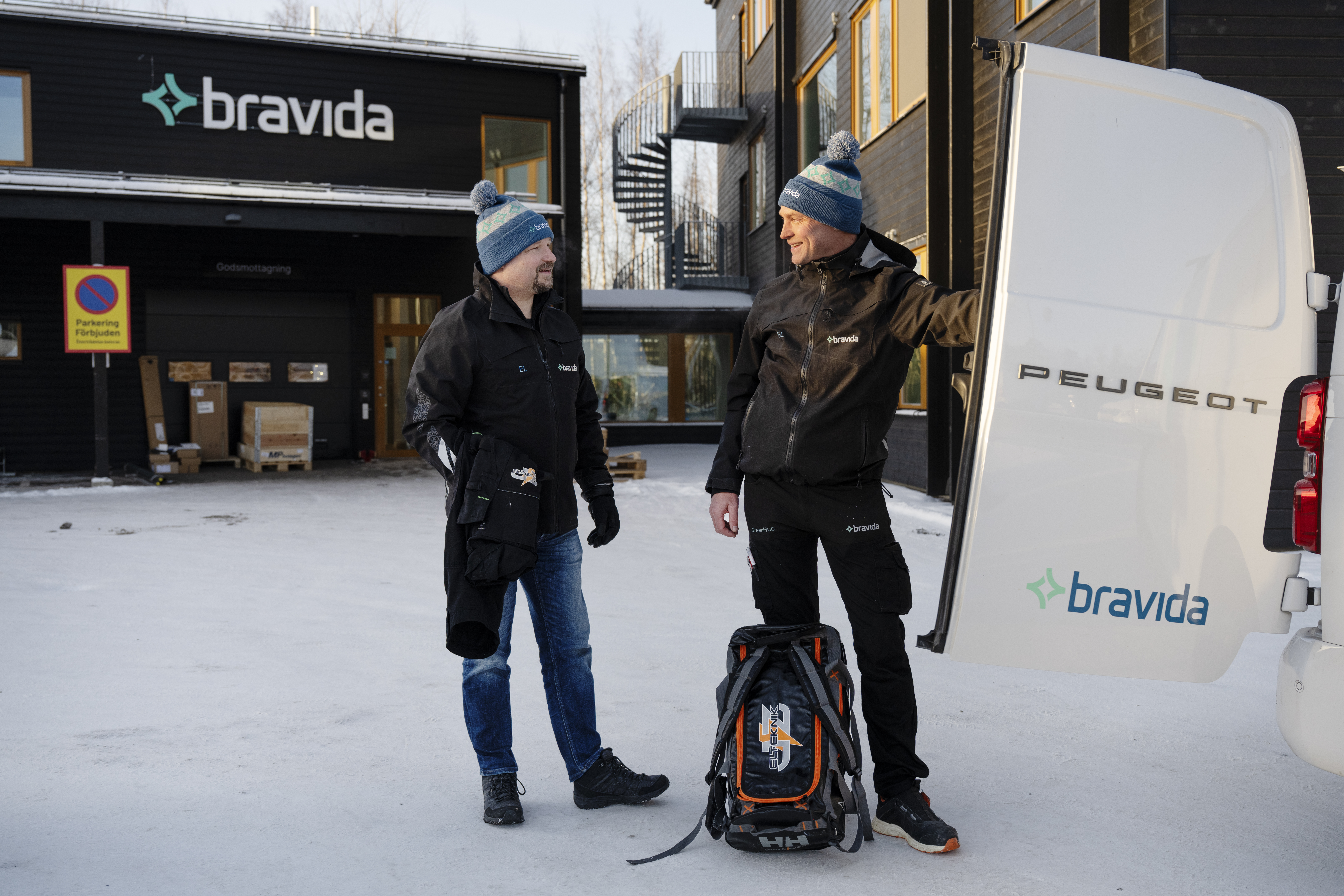 Två män i vinterjackor och mössor står vid en vit Peugeot-skåpbil utanför en Bravida-byggnad med ryggsäckar och utrustning på snötäckt mark.