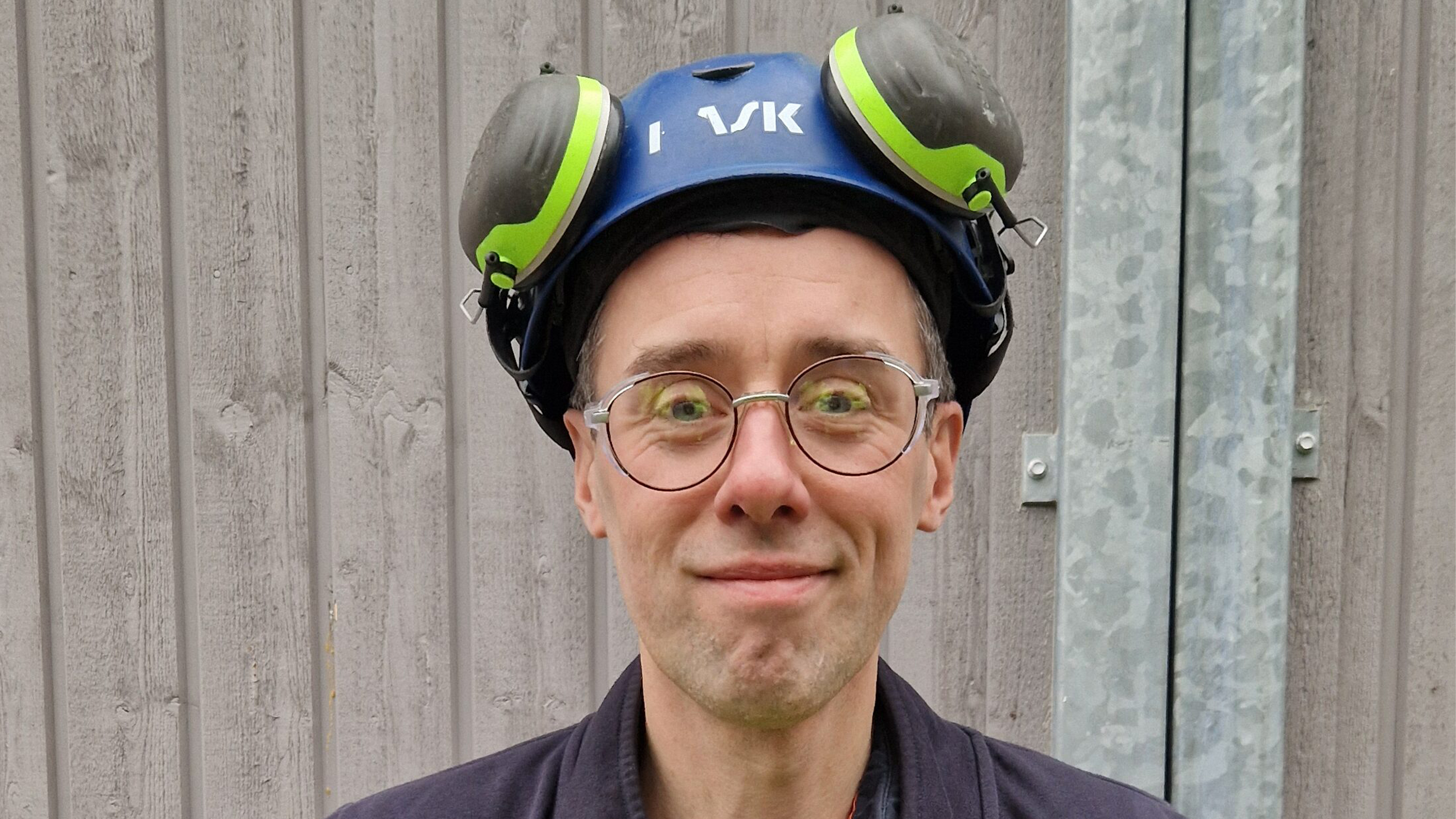 En person med glasögon, blå hjälm med hörselskydd och mörk jacka står framför en vägg av trä.