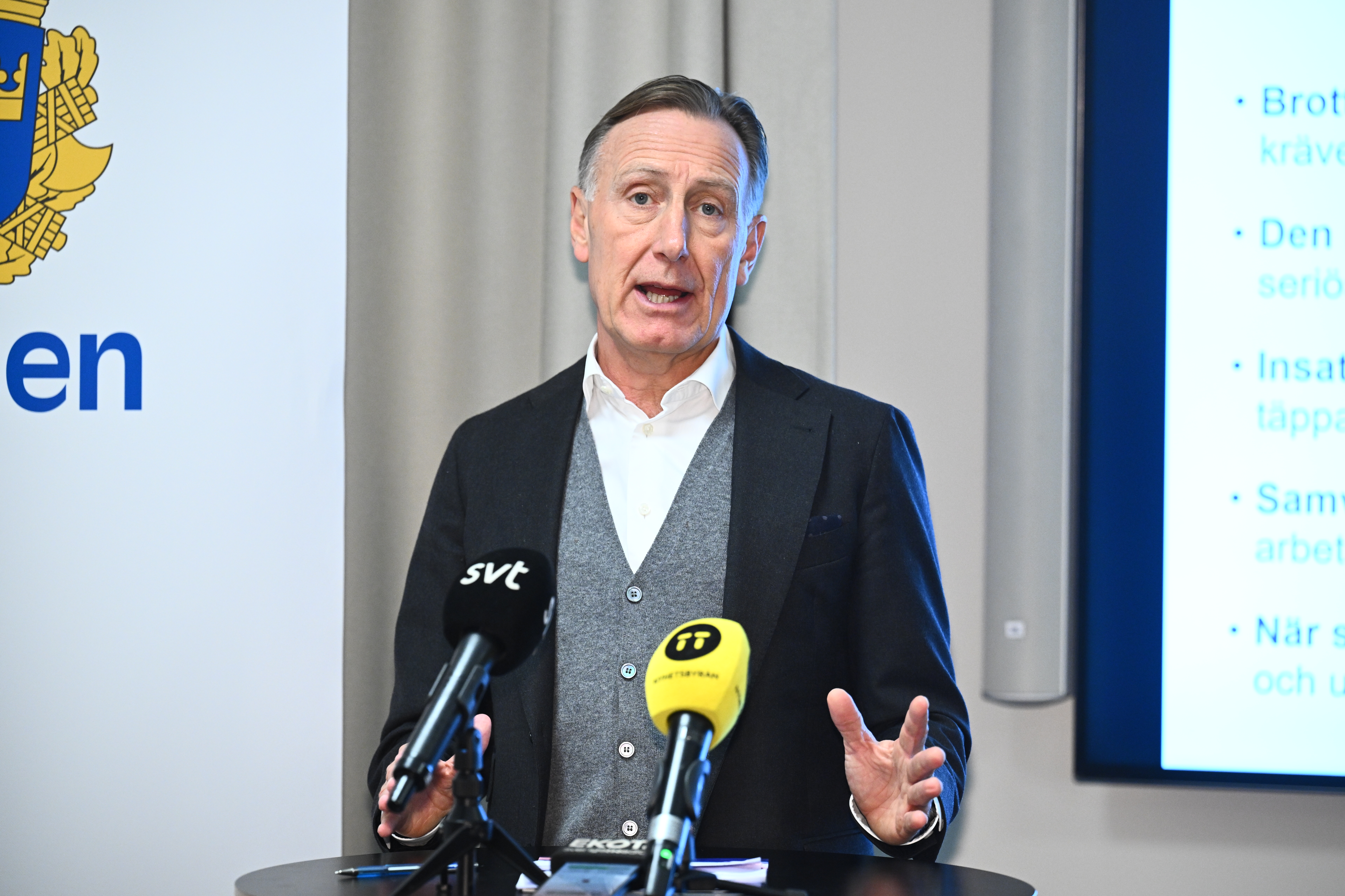En man i kostym talar vid ett podie med mikrofoner framför sig, bredvid en polislogotyp och en skärm med text.