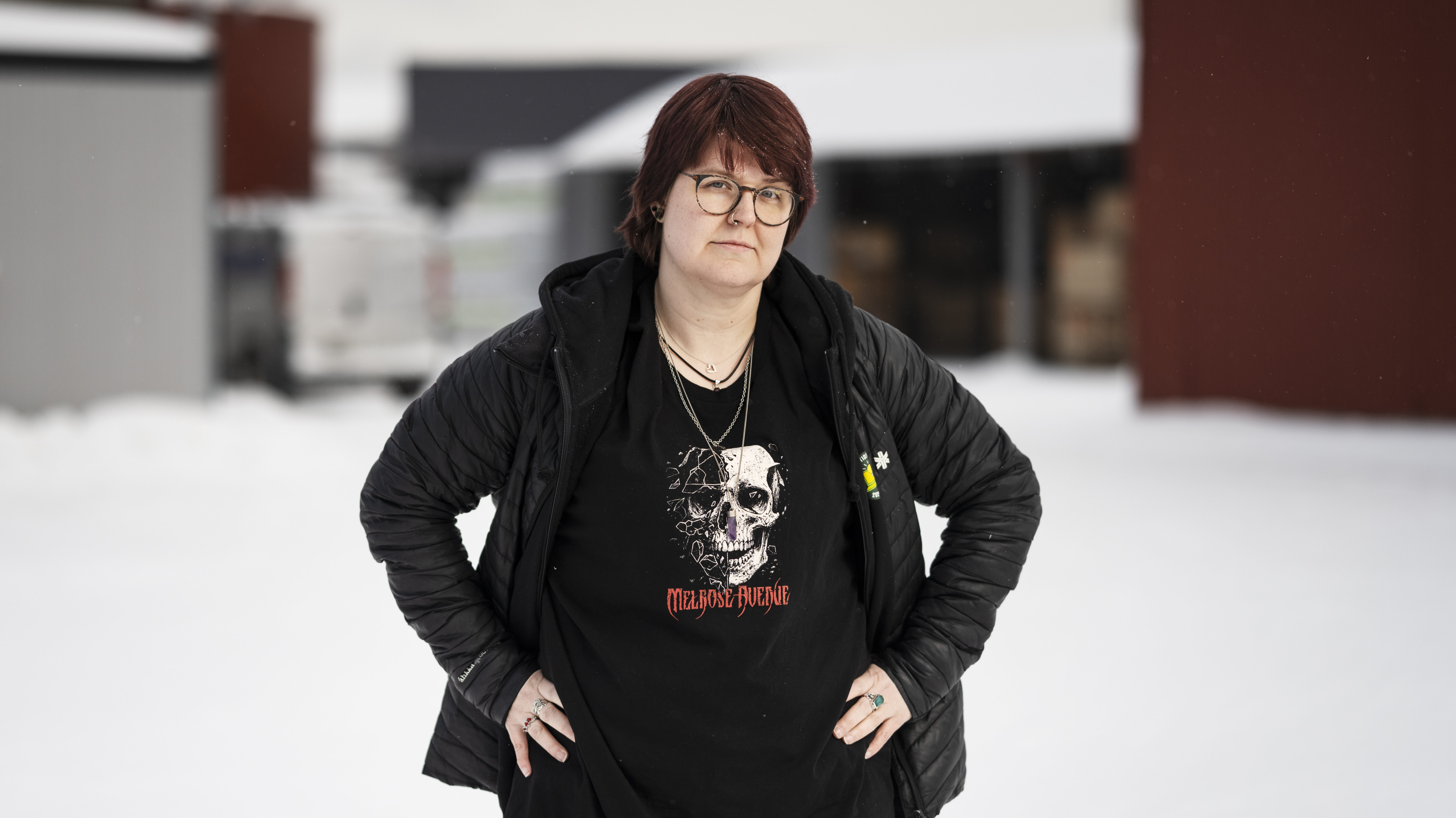 En person med kort brunt hår, glasögon och svart jacka står ute på snö med händerna i sidan, iklädd t-shirt med dödskalle och text.